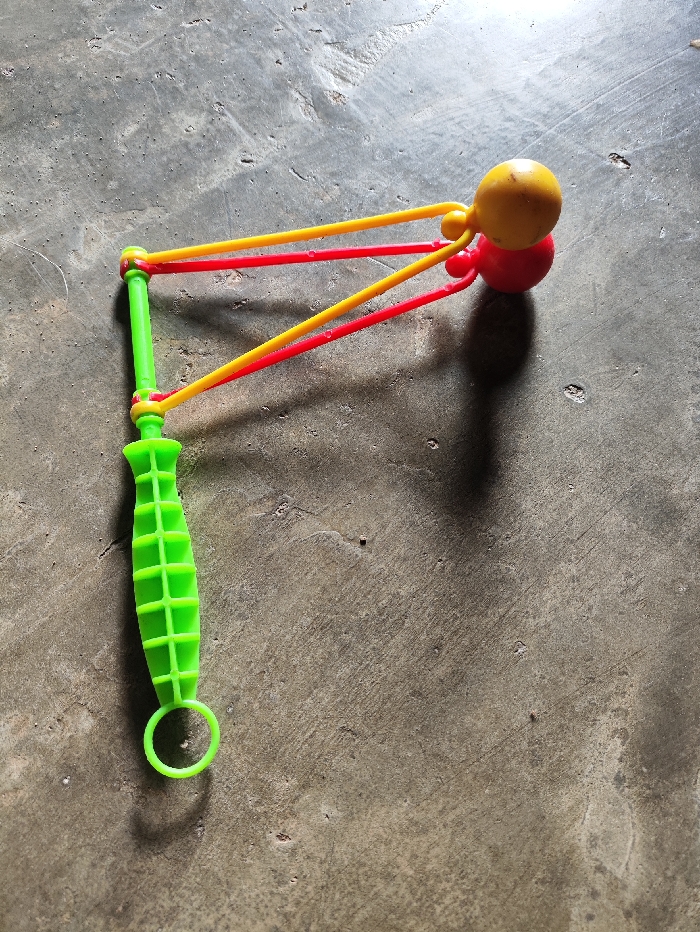 The image captures a brightly colored plastic toy lying on a rough, grey concrete surface. The toy consists of a neon green handle, textured with a ribbed pattern for grip, ending in a small circular loop at its base. Attached to the top of the green handle are two sets of elastic strings or bands: one pair in red and another in yellow. These bands converge to hold two small, spherical objects at their ends—a red sphere at the bottom, topped by a yellow sphere. The design suggests it is a toy slingshot or catapult.
No activity is taking place; the object is static on the ground. A strong, distinct shadow is cast by the toy, indicating bright daylight and sunny weather, likely mid-day or early afternoon. The concrete ground appears weathered and uneven, suggesting an outdoor setting or a simple, unpolished floor. There are no people visible, and no emotions are discernible. No text is present on the toy or in the background. The simple, colorful plastic construction is characteristic of a children's plaything. The location context is Selagai Lingga, Indonesia.