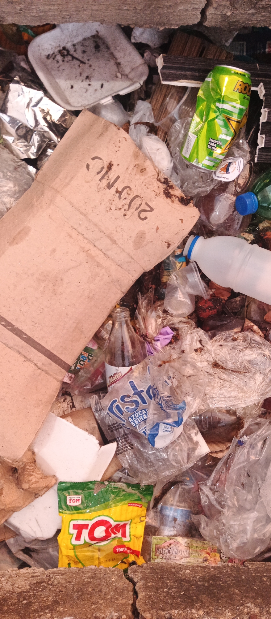 The image shows a close-up, top-down view of a pile of garbage. The garbage includes various items such as crumpled aluminum cans, plastic bottles, a cardboard box, and plastic packaging. Some of the plastic bottles have blue caps. A crumpled green can with the word "ROC" and "ENERGÍA PARA TU DÍA" is visible. A yellow and red packaged item labeled "TOM" is also present, along with a smaller package that says "RICO GRANDE". The garbage is resting on a rough, textured surface that appears to be dirt or soil. The scene suggests an outdoor environment, possibly an alley or a refuse area. There are no people or animals visible in the image. The overall impression is one of discarded waste.