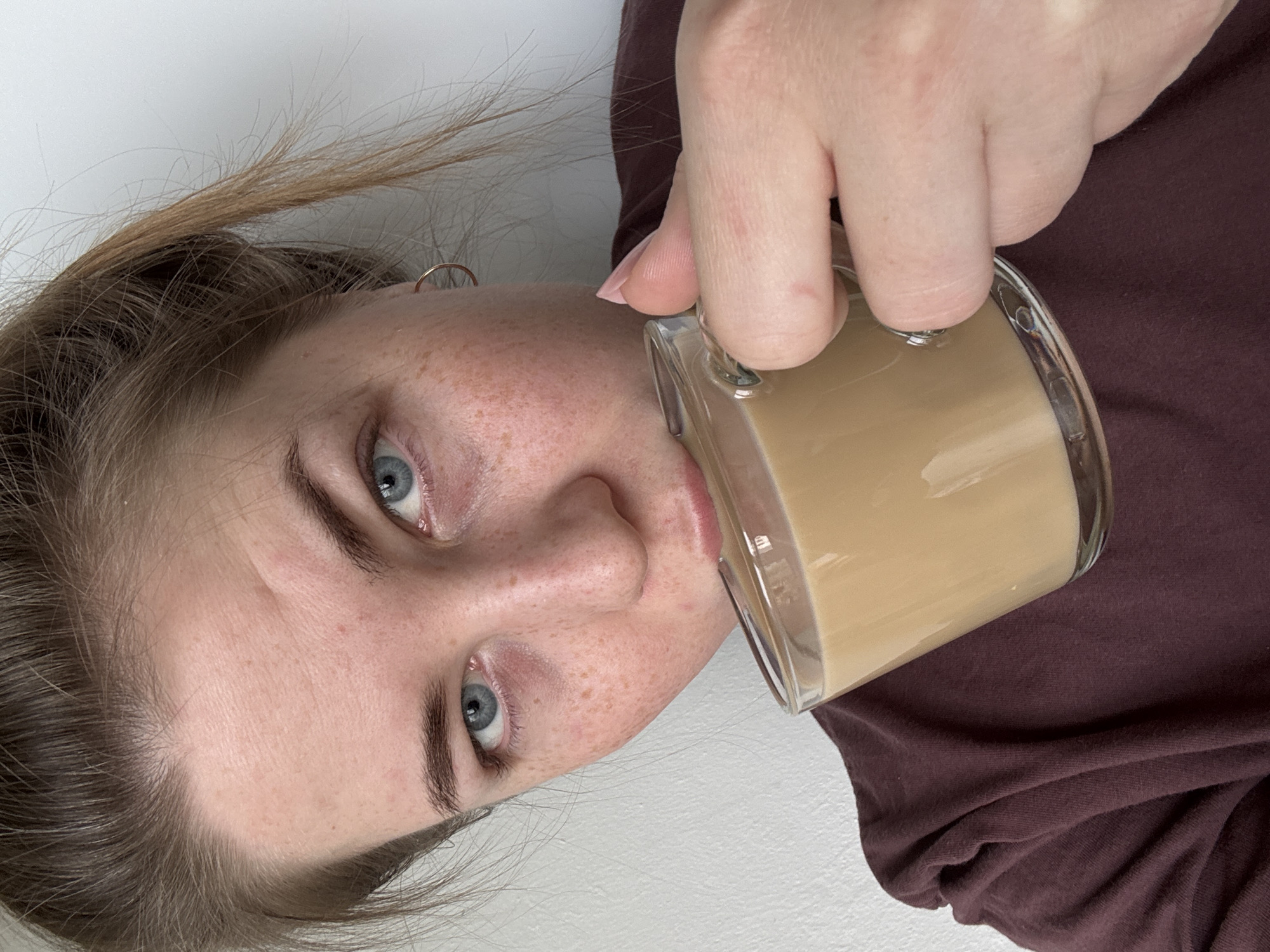 A young woman is captured in a close-up, diagonal shot as she drinks from a clear glass mug in an indoor setting in Chaikovsky, Russia. She has fair skin, prominent freckles across her nose and cheeks, and striking blue eyes. Her light brown hair, which appears slightly disheveled, frames her face. She is wearing a dark red or maroon top, visible on her right side.
The woman is holding a clear glass mug with a handle, filled with a light beige or creamy brown beverage, likely coffee or tea with milk, to her lips. Her left hand is visible, gripping the mug. A small gold hoop earring is visible on her left ear. Her expression is neutral, focused on the act of drinking. The background is a plain, light-colored, likely white, wall, suggesting a simple, casual environment. There is no visible text in the image.