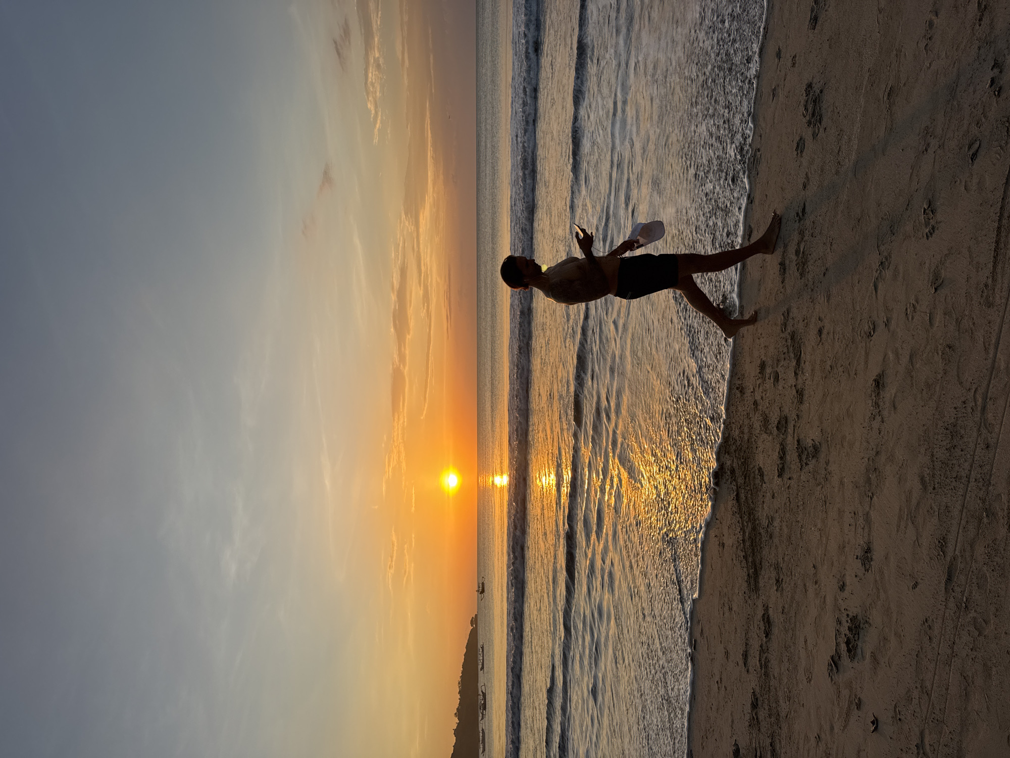 A silhouetted figure stands on a sandy beach at sunset in Kuta Selatan, Indonesia. The person holds a phone in one hand and a white object in the other, partially submerged in the receding surf. The sunset casts a golden glow across the water and sky. The sky is gradient, fading from orange to gray. The sea has gentle waves approaching the shore. The sand shows footprints. In the distance, silhouetted boats are visible.