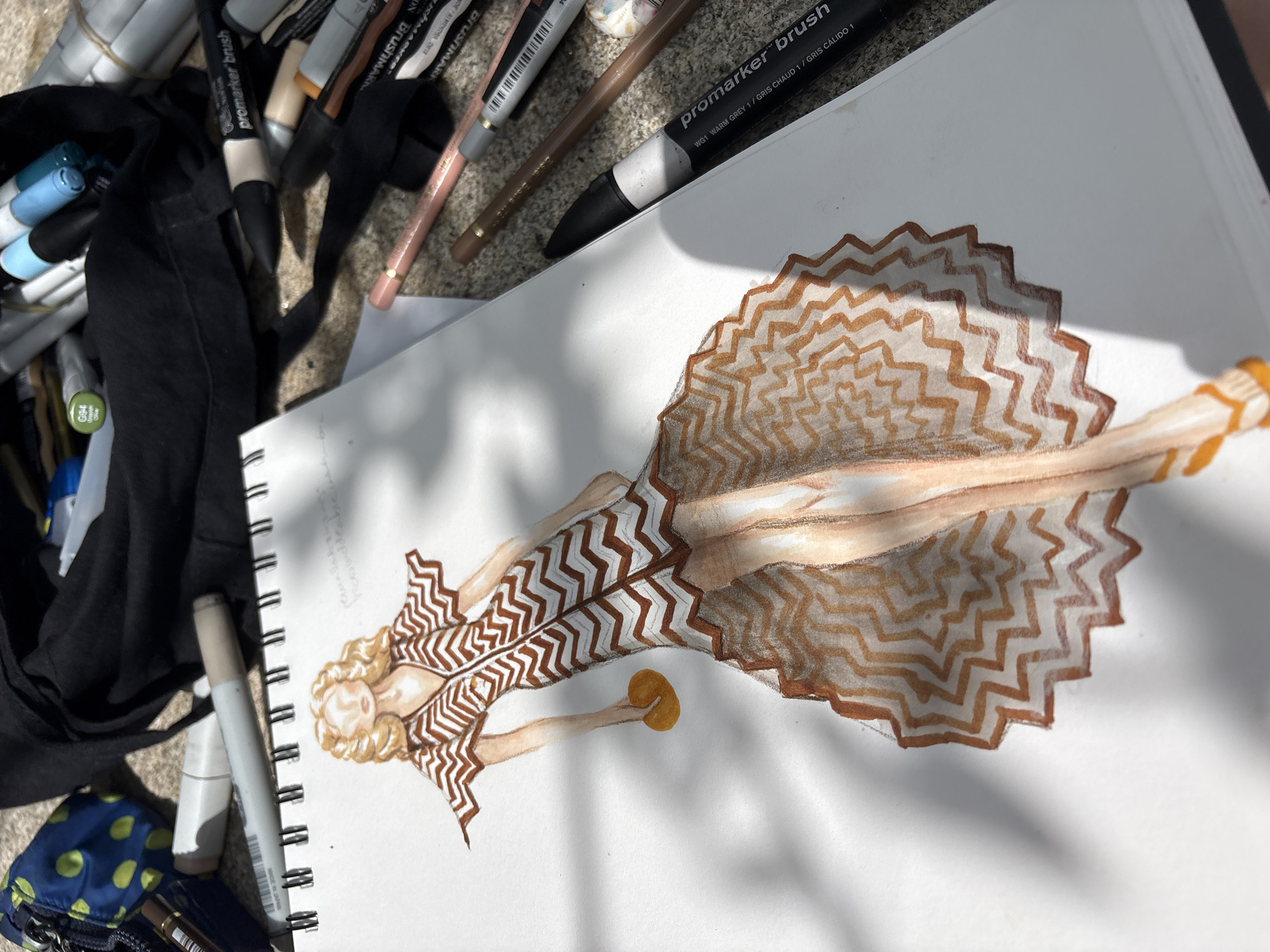 A fashion sketch of a woman in a zigzag patterned dress is depicted on a white sketchbook page. The sketch is done with colored pencils and markers. The woman has blonde curly hair and is holding a round, yellow object in her left hand. Her dress has a V-neck and short sleeves, with a flared, floor-length skirt. Orange sandals are visible on her feet.
The sketchbook is open on a textured surface, possibly concrete or stone, with dappled sunlight casting shadows. Surrounding the sketchbook are various art supplies, including colored pencils, markers, and a black fabric pouch. Some of the markers are Promarker Brush pens, and one is labeled with "G94" and "Olive". The art supplies are scattered around the sketchbook, suggesting an outdoor creative session. The image does not provide any specific information about the location in the city of O Casal, Spain.
