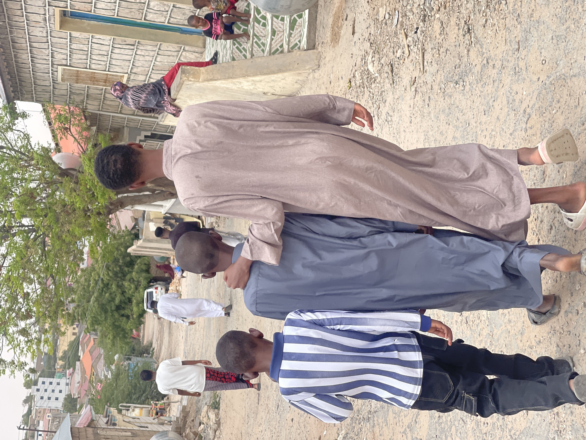 In Hargeisa, Somaliland, a group of boys walks down a dusty, unpaved street lined with buildings. The architecture suggests a blend of traditional and modern styles, with some structures appearing to be made of concrete and corrugated metal, while others are more elaborately designed with stone facades. The road is uneven and stony, indicative of unpaved urban infrastructure common in many parts of the city.
In the foreground, three boys are walking away from the camera. The boy on the left is wearing a blue and white striped long-sleeved shirt and dark cargo pants. The middle boy is in a dark blue thobe. The boy on the right, the tallest of the three, is wearing a light brown thobe. He has his arm around the shoulders of the middle boy. Further down the street, two other boys are visible walking towards the camera, one in a white thobe and the other in a white t-shirt and patterned pants. In the background, more people are seen, including a woman in a colorful headscarf and red pants sitting on a low wall, and two young children sitting on steps. A white SUV is parked on the side of the road. The surrounding environment is characterized by residential buildings, some with multi-story designs, and sparse vegetation, typical of an urban setting in a semi-arid climate. The overall impression is of a community going about its daily life in a bustling, developing urban area.