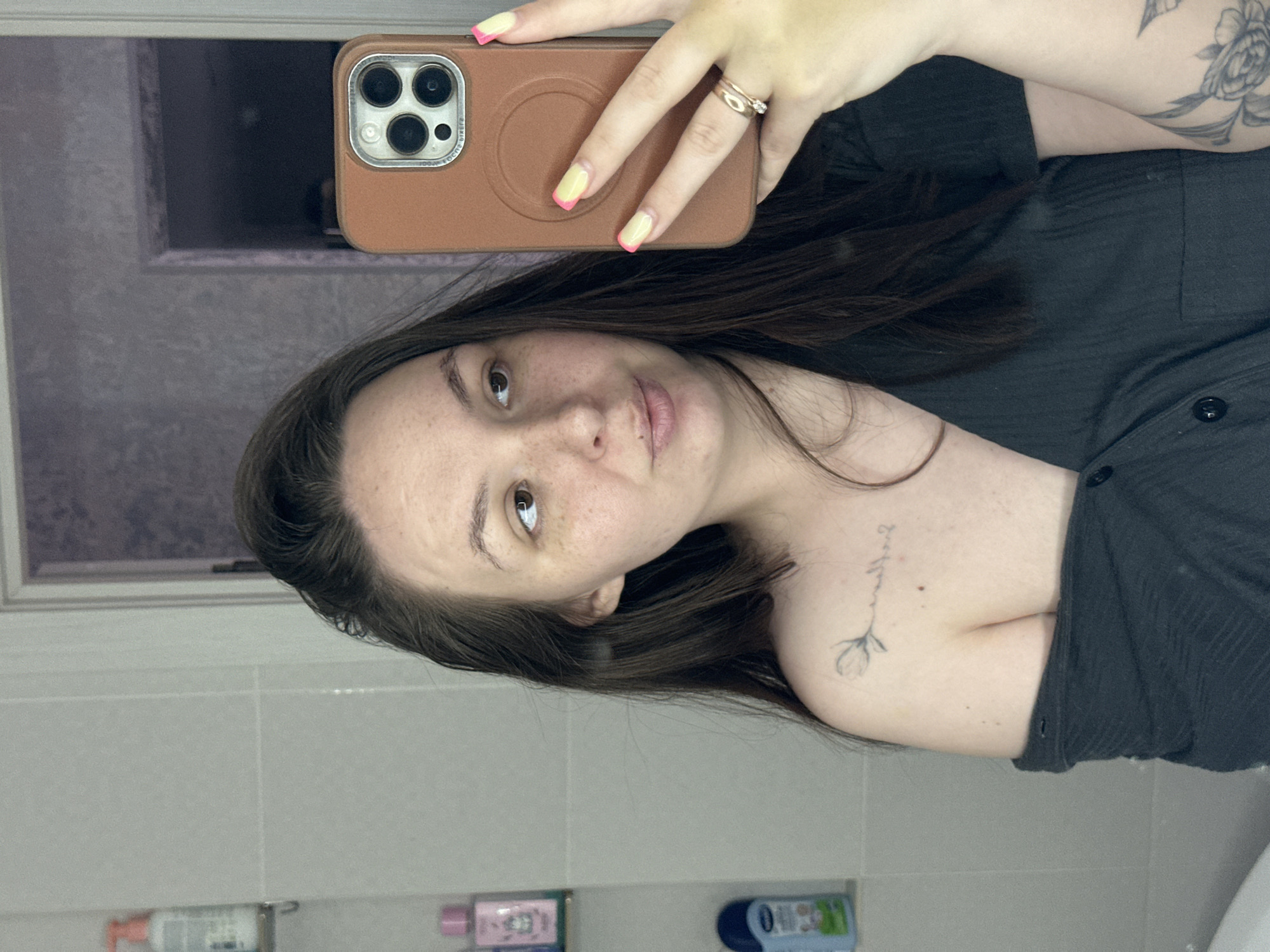A young woman with long, dark hair, freckles, and light brown eyes is taking a selfie in a bathroom. She is wearing a dark, off-the-shoulder top. She has a small tattoo of a flower on her left shoulder and a larger floral tattoo on her right arm, partially visible. She is wearing two rings on her right ring finger. Her fingernails are painted with a yellow base and pink tips. The background shows a tiled wall, shelves with toiletries, and a closed door. This image was taken in Chaikovsky, Russia.