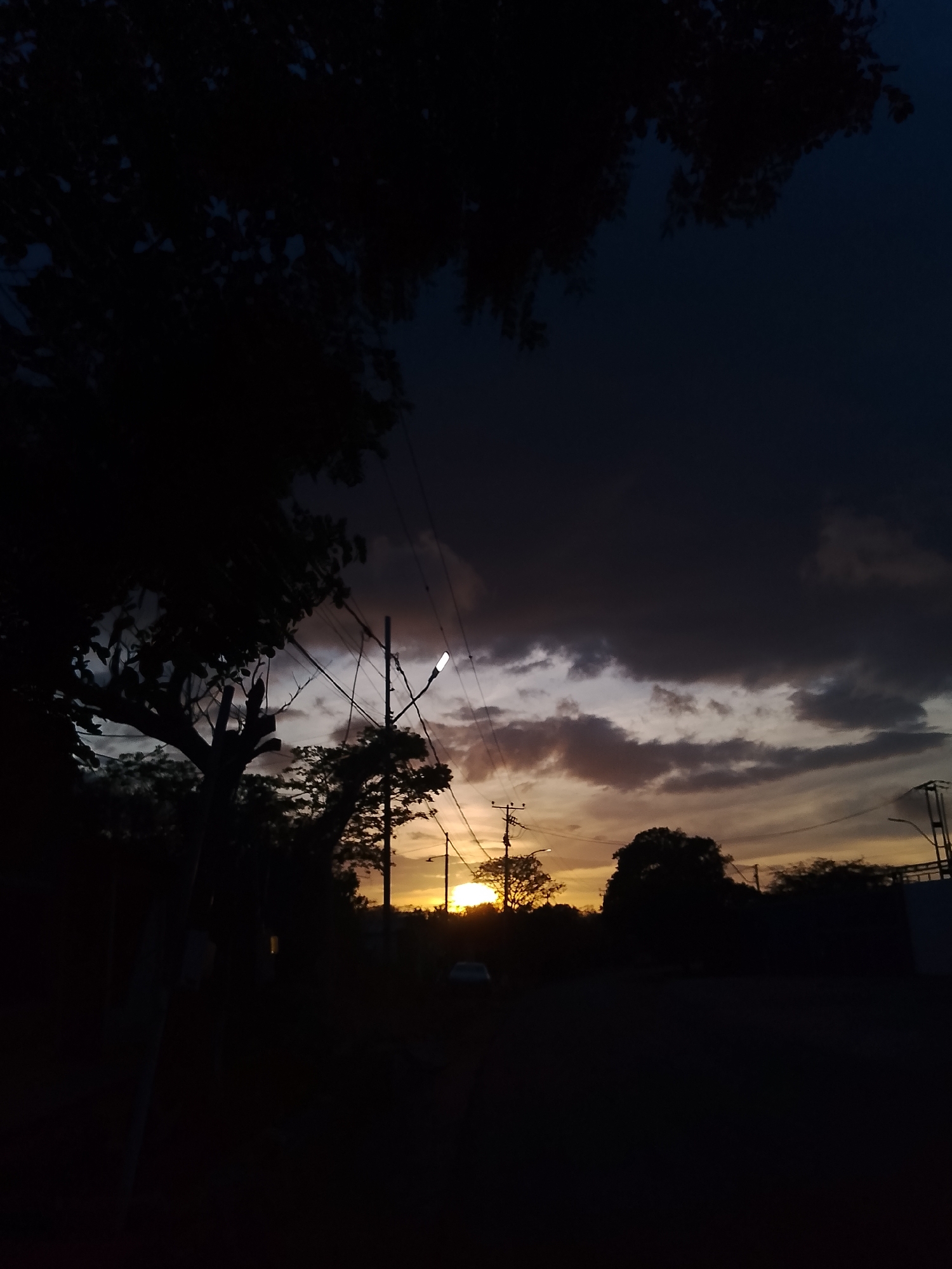 The image depicts a street scene at dusk in Santa Ana, Venezuela. The sky is filled with dark, heavy clouds tinged with hues of orange and yellow from the setting sun. Silhouetted against the sky are several trees, utility poles with visible wires, and a streetlamp that is lit. The foreground is dominated by the dark silhouette of a large tree on the left and a street or road. A faint outline of a white car can be seen in the middle ground. The overall impression is one of a quiet evening in a residential or semi-rural area of the city.