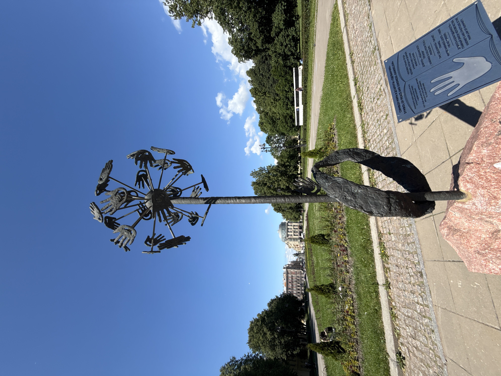 This is a photograph of a sculpture in Saint Petersburg, Russia, set against a clear blue sky with scattered white clouds. The sculpture, located on a paved area with a pink granite base, is made of dark metal and depicts a dandelion-like structure with numerous hands extending outwards from the central stem. Below the sculpture, a dark blue plaque with white text and an illustration of a hand is partially visible. The background features a well-maintained grassy area with small evergreen bushes and trees. Beyond the green space, there are buildings with light-colored facades and a distant white bus. The overall scene is one of an outdoor public space under bright daylight.