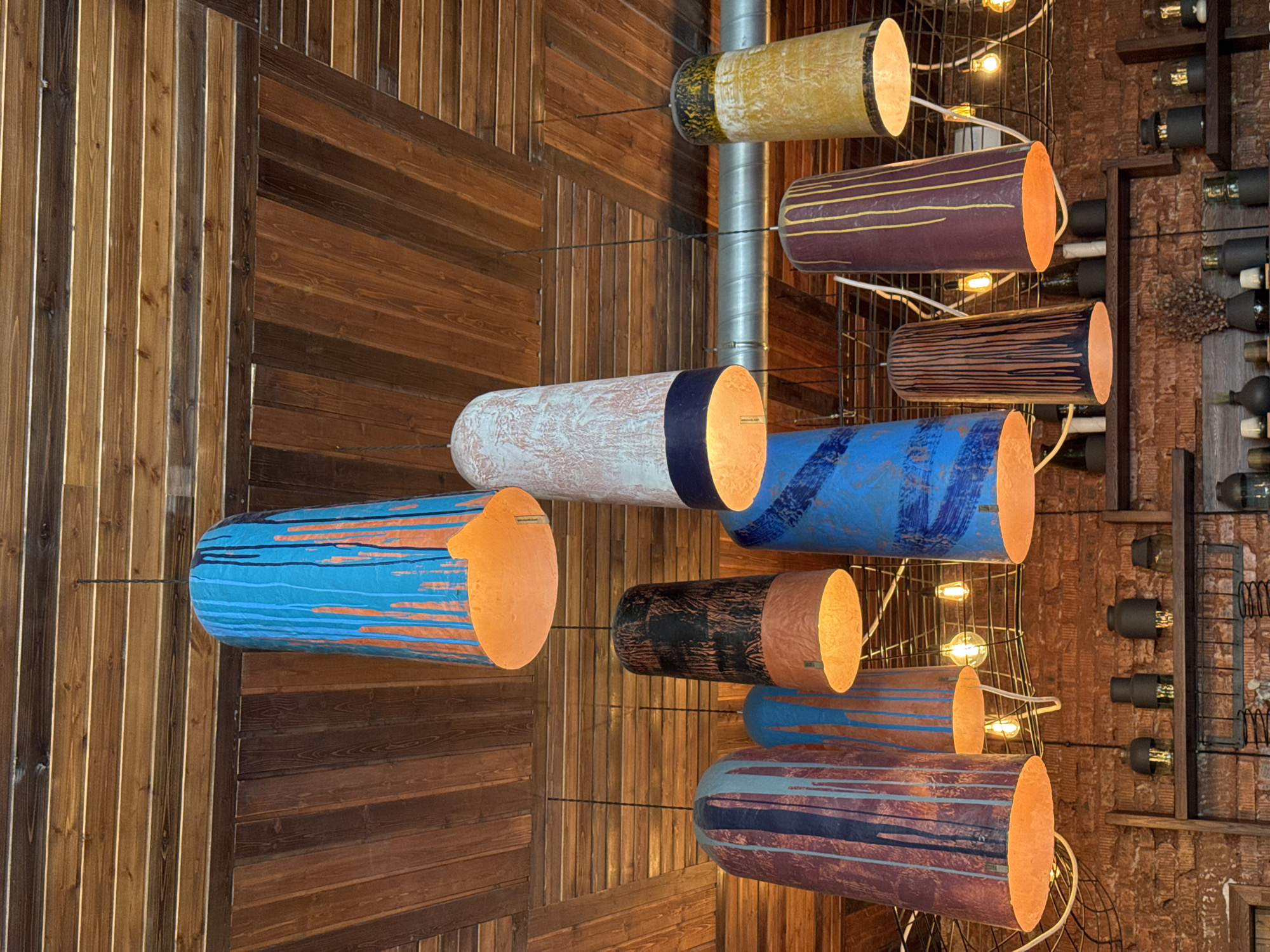 This media file presents an indoor scene, likely from a restaurant, bar, or unique commercial space in Birobidzhan, Russia, characterized by its distinctive lighting and extensive use of wood. The primary subjects are numerous cylindrical pendant light fixtures suspended at various heights and angles, dominating the center of the image.
These lamps are varied in size and are decorated with abstract, hand-painted patterns featuring vibrant colors such as blues, oranges, purples, yellows, and earthy browns, often with contrasting stripes or distressed textures. The interiors of the cylinders emit a warm, inviting orange glow, suggesting incandescent light sources. Thin black wires support each fixture, creating a sense of them floating. A prominent silver metal ventilation pipe runs vertically in the background on the right, partially integrated with the lighting arrangement.
The background consists of rich, warm-toned wooden paneling that covers both the walls and ceiling. The wood grain patterns vary, with sections arranged vertically, horizontally, and diagonally, adding visual depth and texture. On the far right, a rustic wooden shelving unit, serving as a wine rack, is visible, filled with numerous dark-colored bottles. Additional bare Edison-style light bulbs are interspersed among the larger cylindrical fixtures, contributing to the overall warm ambiance.
No people are visible in the scene, which focuses entirely on the interior design and lighting. The illuminated lamps and warm wood tones create a cozy and inviting atmosphere, suggesting it is either evening or a dimly lit interior. On several of the light fixtures, the text "smac.ru" is visible, along with some smaller, less legible text beside it.