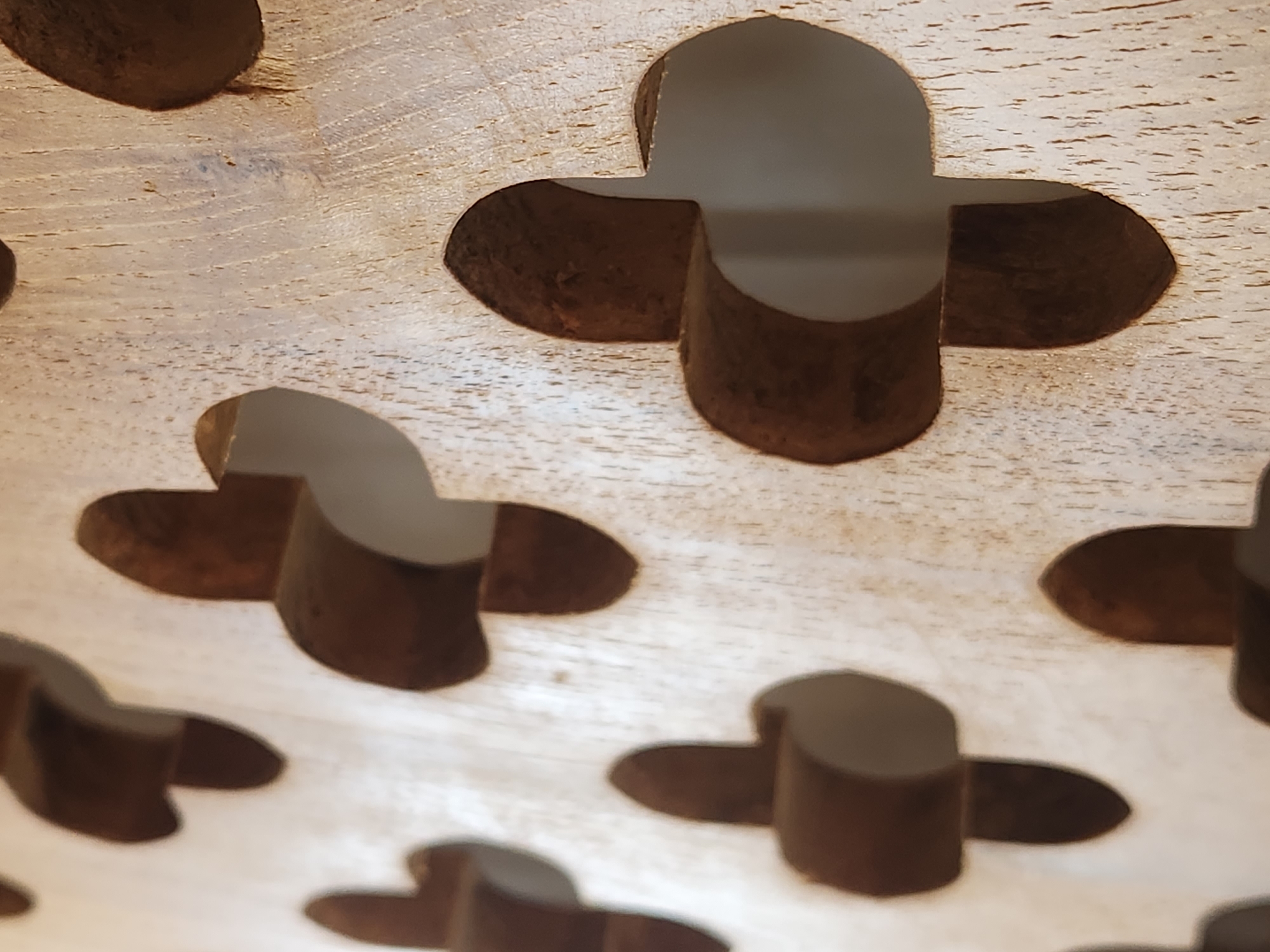 The image shows a close-up, angled view of a wooden screen with a repeating clover-like pattern cut out. The wood appears light-colored with a visible grain and some imperfections. The cut-out shapes are dark brown and three-dimensional, suggesting they are carved or molded elements within the screen. The background visible through the cut-outs is a muted, off-white color, possibly a wall or another surface. The lighting casts subtle shadows within the carved spaces, highlighting the depth and texture of the material. The scene is devoid of any people or specific identifiable objects that would indicate a location within the city of Maitland, United States of America.
