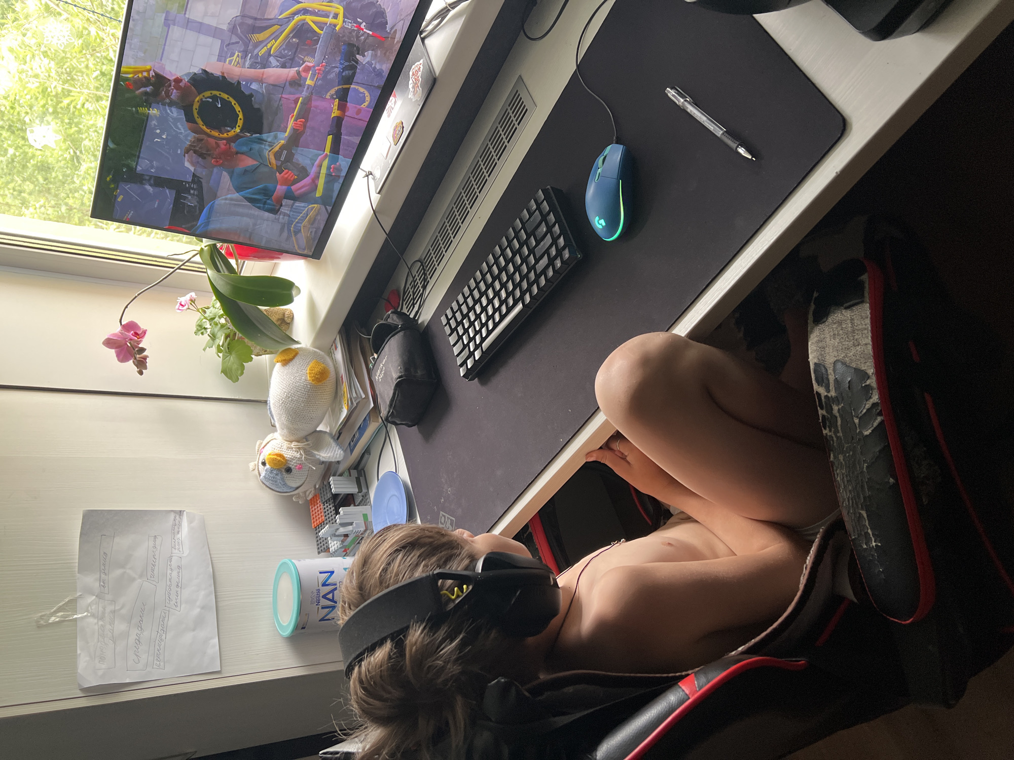 The image depicts a child sitting in a gaming chair, wearing headphones and possibly engaged in playing a video game on a computer. The computer setup includes a monitor displaying a game, a keyboard, a mouse, and a mousepad. The desk also has a pen, a can of Nestle NAN, and a sheet of paper with handwritten text. An orchid and a stuffed toy are visible next to the monitor. The chair appears worn. The city name is not mentioned in the image. The OCR data seems to be from a document on the desk.