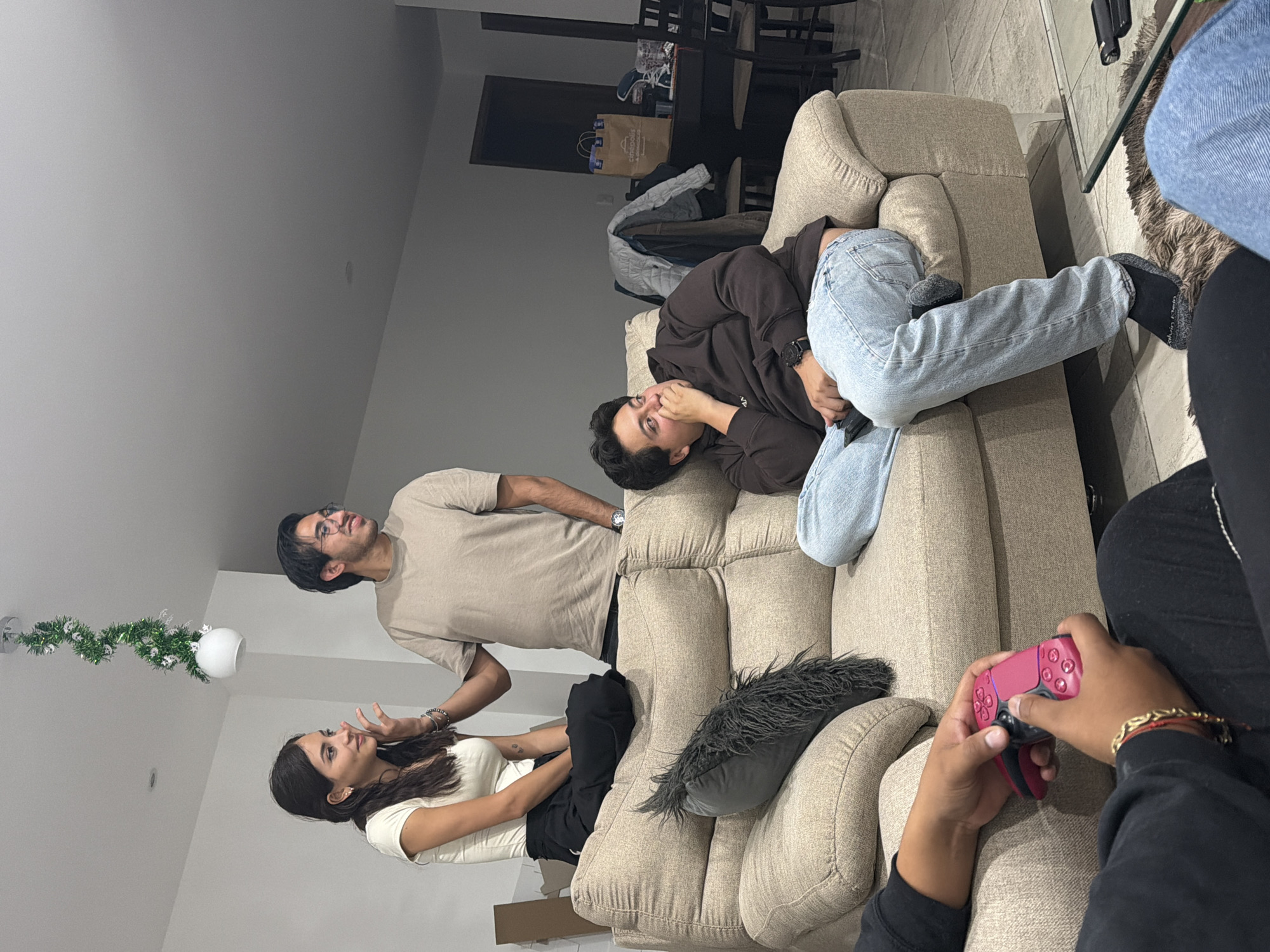 The image shows a group of four people gathered on a beige sectional sofa in what appears to be a living room. A woman in a white shirt is seated at the end of the couch, gesturing with her hands while talking to a man in a tan shirt who is standing beside her, leaning over the back of the sofa. Another man wearing a brown hoodie and light blue jeans is lying on the couch, looking at the camera. A fourth person is partially visible, seated on the couch, holding a pink gaming controller. A decorative garland is attached to a light fixture on the ceiling. In the background, there is a dining table with chairs, and a door leading to another room. A "Cinepolis" branded shopping bag is on the table. The location is San Andrés Cholula, Mexico.