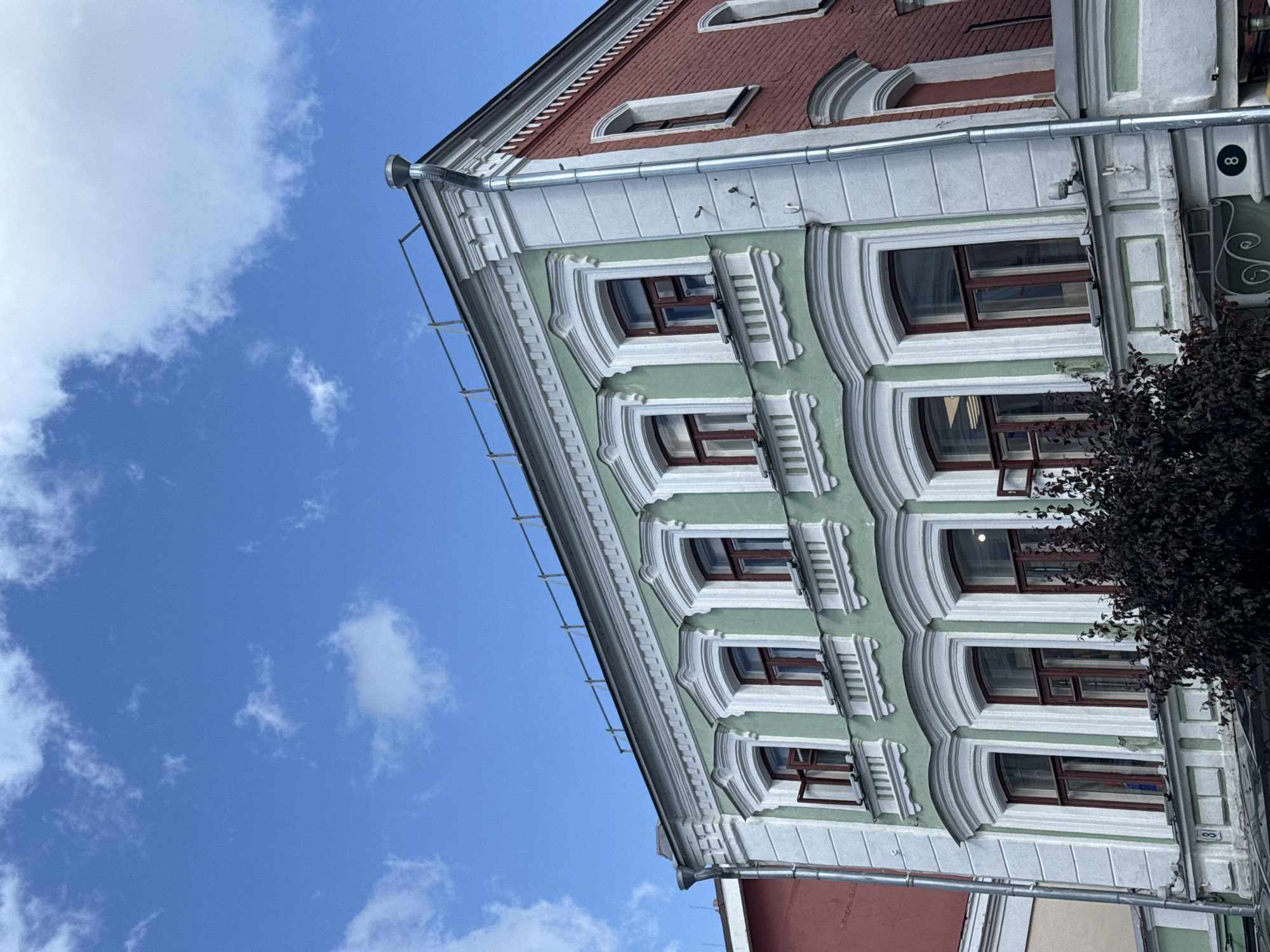 The image captures an upward view of an old building with two distinct facades: one painted in a pale green and the other in red brick. The green section of the building features several windows with white, ornate trim, and dark wooden frames, some of which are partially open. The red brick section also has windows with arched tops and white trim. A metal downspout runs vertically along the corner where the two facades meet. The building has a detailed cornice and a railing along its roofline. Below the windows of the green facade, a dark, leafy bush is visible. In the bottom right corner of the image, a circular sign with the number "8" is present. The building is situated under a bright blue sky with scattered white clouds. The architectural style suggests it is likely located in the historic center of Nizhny Novgorod, Russia, given the common style of buildings found in that area.