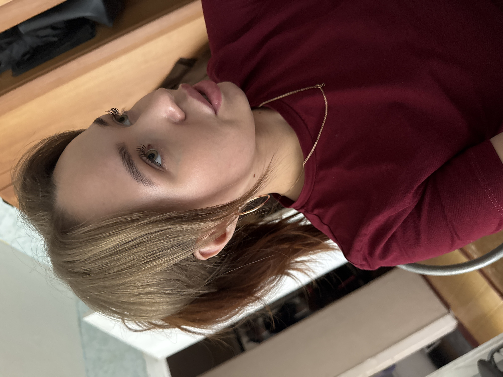 A young woman with auburn hair pulled into a ponytail and wearing a maroon t-shirt and a gold chain necklace is looking to the right of the frame. She has on large hoop earrings. The background consists of a wooden closet and a doorway with a light-colored wall and wooden floor. The lighting suggests an indoor setting, likely a home, in the city of Chaikovsky, Russia.