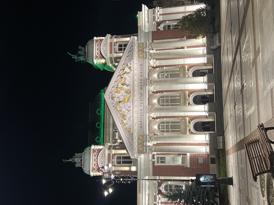 This image depicts the Ivan Vazov National Theatre in Sofia, Bulgaria, at night. The grand Neoclassical building features a red and white facade with prominent white Corinthian columns and ornate gold architectural details, including a sculptural frieze and pediment above the central entrance. The building's main facade is illuminated by numerous upward-facing white spotlights. Two towers flank the central structure, each topped with sculptural groups; the right tower features a quadriga-like sculpture. The theatre's roof and parts of its domes glow with a distinct green light. The name "НАРОДЕН ТЕАТЪР - ИВАН ВАЗОВ" is prominently displayed in gold lettering above the main entrance. In the foreground, an expansive, empty paved plaza extends from the theatre steps, featuring a pattern of light and dark rectangular tiles. A dark wooden slatted bench with black metal supports is positioned in the lower-left corner, facing the theatre. To the left, a black, four-lamp street light stands next to a dark conifer tree and a vertical poster display. Another conifer is visible on the right side of the theatre. The sky is dark, indicating nighttime conditions. No individuals are visible within the frame.