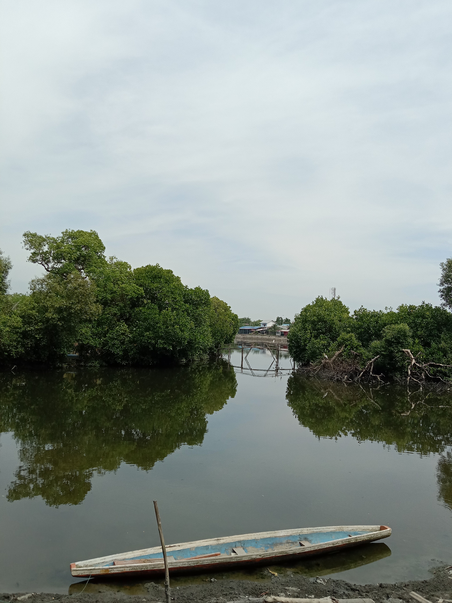 The image shows a still body of water, likely a river or inlet, flanked by dense green vegetation on both sides, reflecting in the water's surface. A small, narrow boat rests on the muddy bank in the lower part of the frame. A simple wooden bridge crosses the water in the distance. In the background, there are glimpses of buildings and a cellular tower, suggesting the presence of a settlement nearby. The overcast sky provides soft, diffused lighting. The scene is located in Lhokseumawe, Indonesia, evidenced by the specific appearance of the vegetation and the architectural style of the distant buildings.