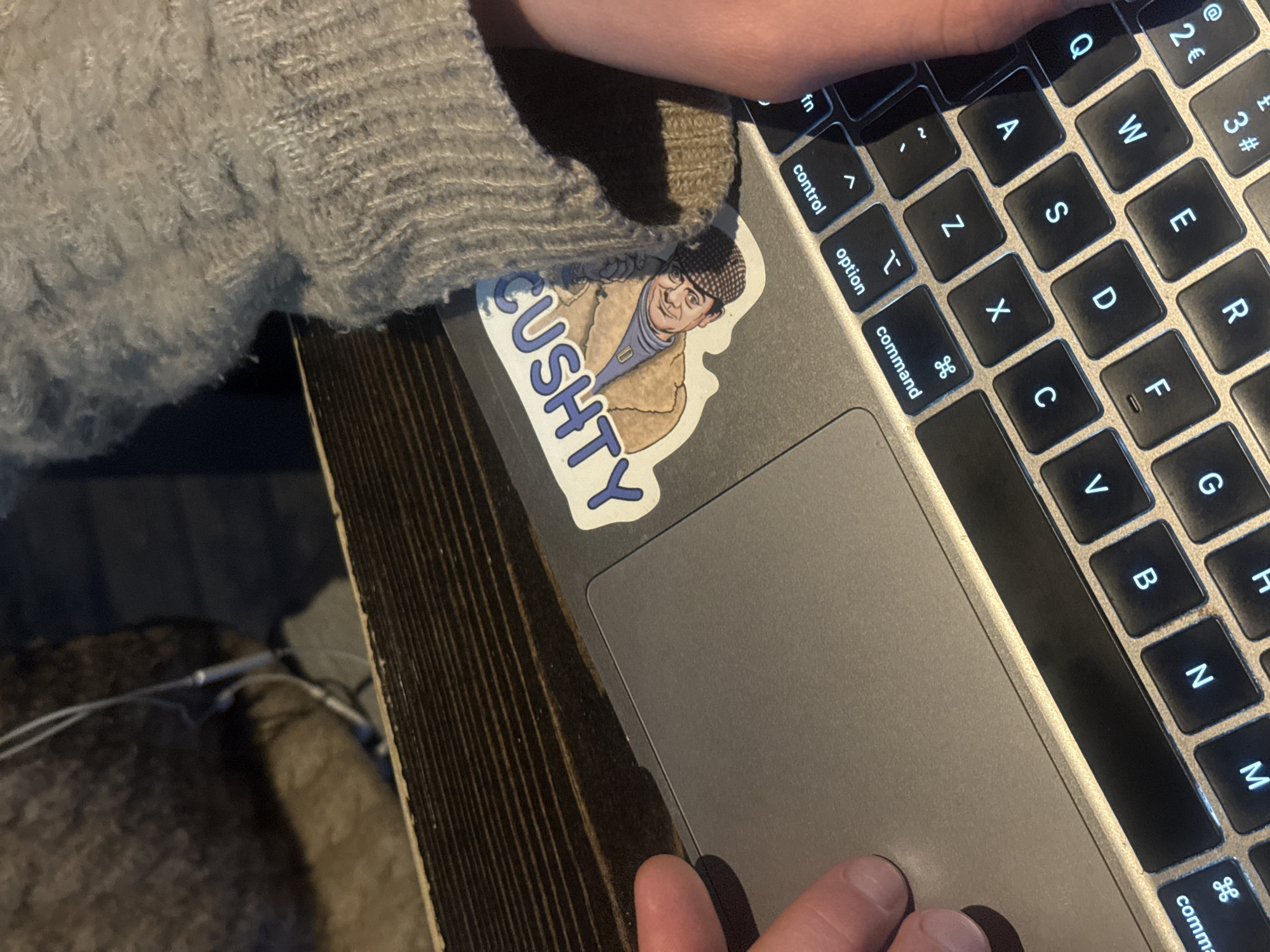 An overhead, angled shot depicts a person's hands operating a silver laptop, likely a MacBook Pro, on a dark wooden surface in London, United Kingdom. The person wears a light grey, fuzzy knitted sweater, with a sleeve partially covering the top left of the laptop.
One hand rests on the trackpad, while fingers of the other hand are positioned over the backlit keyboard. The keyboard shows a European layout with keys such as Q, W, E, R, A, S, D, F, G, H, Z, X, C, V, B, N, M, fn, control, option, command, comma, 2€, and 3#.
A prominent sticker is affixed to the laptop's casing, left of the trackpad. It features a smiling cartoon man in a checkered flat cap, a beige coat, and a blue turtleneck. Beside him, the word "CUSHTY" is written vertically in bold blue letters with a white outline.
The scene is indoors, suggesting a desk or table setup. Blurred dark objects and thin white wires are visible in the background. The laptop's keyboard backlight is active, suggesting dim ambient lighting. The image offers a close-up view of a person engaged in computer use.