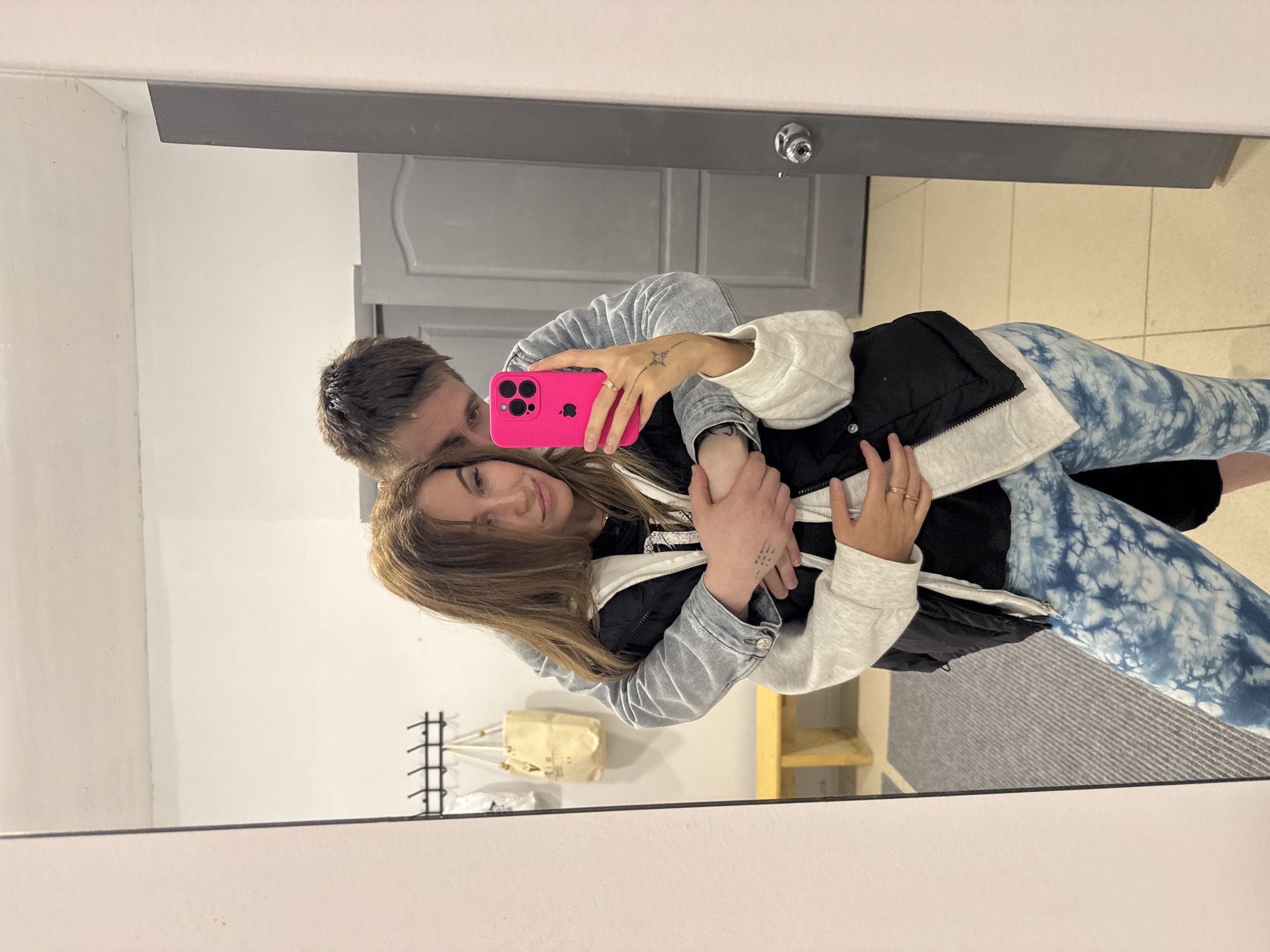A couple is posing for a selfie in a dressing room. The woman is holding a pink smartphone with multiple camera lenses. She has blonde hair and is wearing a black vest over a white hoodie, blue tie-dye pants, and a ring on her left hand. The man has short brown hair and is wearing a denim jacket. They are embracing, and the man's hand is over the woman's hand, which holds the phone.
The background shows the interior of a dressing room with light-colored walls and a mirror. A clothes rack with a white bag hangs on the left side of the frame. The floor appears to be tiled. A door frame is visible behind them.
There is no information within the image to determine it was taken in Chaikovsky, Russia.