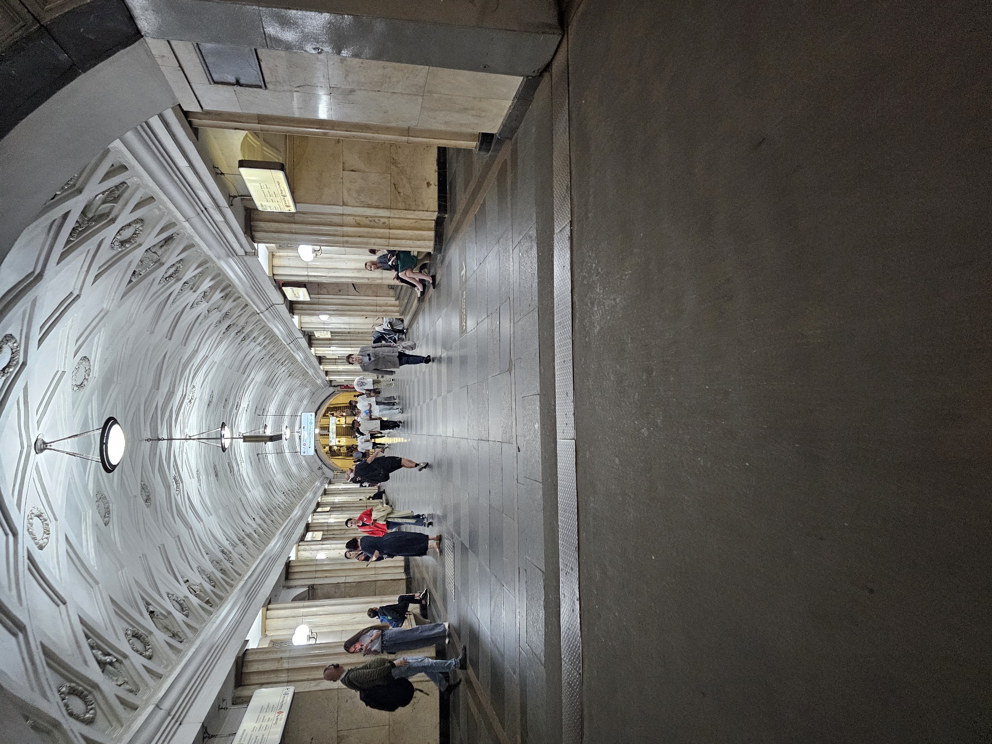 This is an image of a metro station in Moscow, Russia. The station features a grand, vaulted ceiling adorned with ornate stucco work and decorative elements, illuminated by hanging light fixtures. The walls are made of marble and stone, with large pillars supporting the arched structure. The floor is tiled with large, dark stone slabs.
Several people are visible in the image, walking along the platform or waiting. They are dressed in casual clothing. Some individuals are carrying bags or backpacks, suggesting they are commuters. Signs with directional arrows and text are mounted on the walls. The overall atmosphere appears to be that of a busy public transportation hub. The perspective suggests the photo was taken from the platform looking down the length of the station concourse.