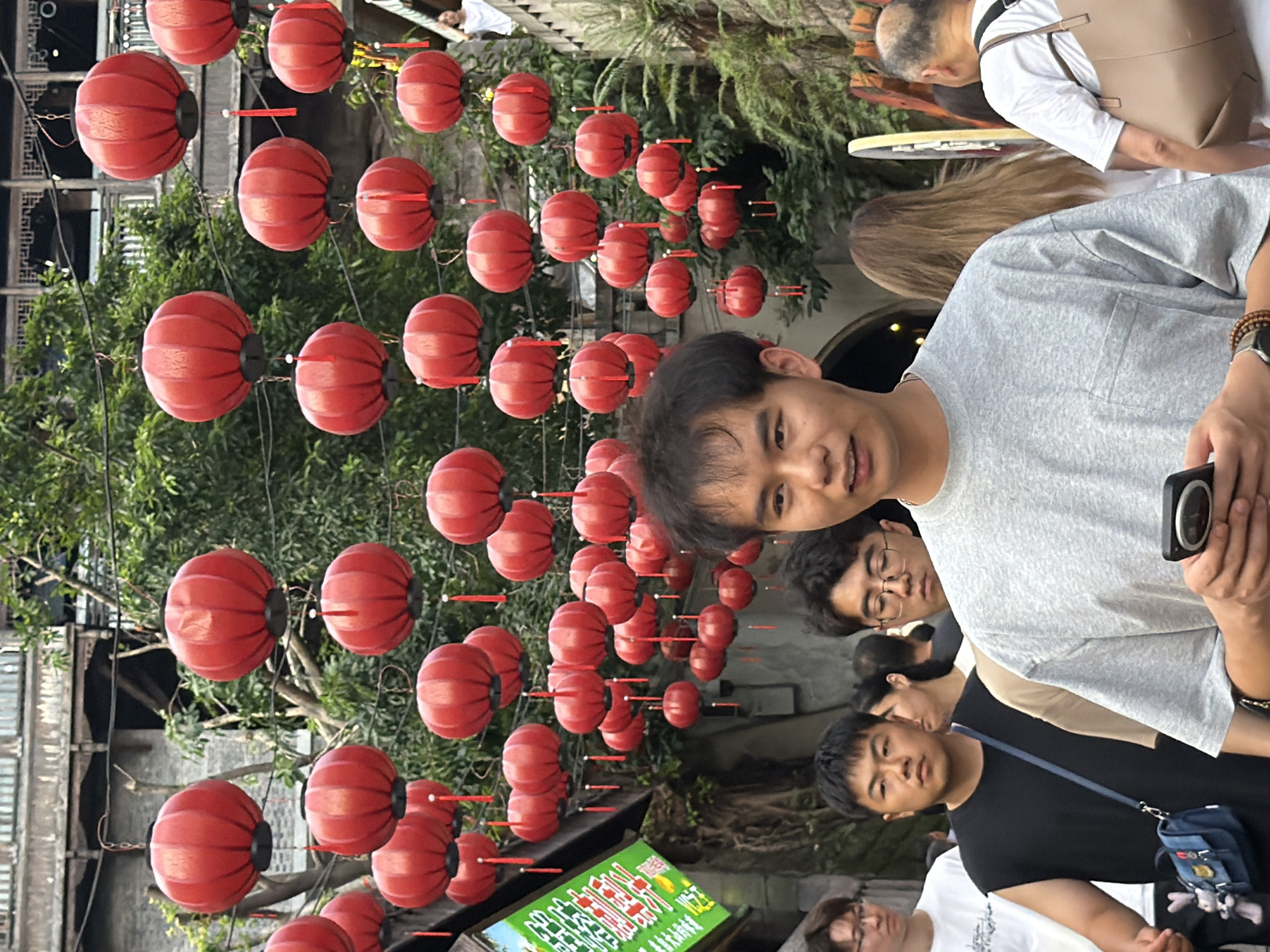 A young Asian man in a grey t-shirt is holding a smartphone in his right hand, looking forward with a slight smile. He is standing in a street scene filled with red lanterns hanging overhead, indicative of a cultural festival or market. The background shows traditional architecture with multiple levels and a lush green canopy of trees. Several other people are visible, including men and women walking and standing around him. One man in a black sleeveless shirt with a blue crossbody bag is to his left, and another man wearing glasses and a white t-shirt is further left. The street appears to be paved, and a sign with Chinese characters is visible on the left side of the frame. This scene is likely in Yuzhong District, China, known for its historical and cultural attractions.