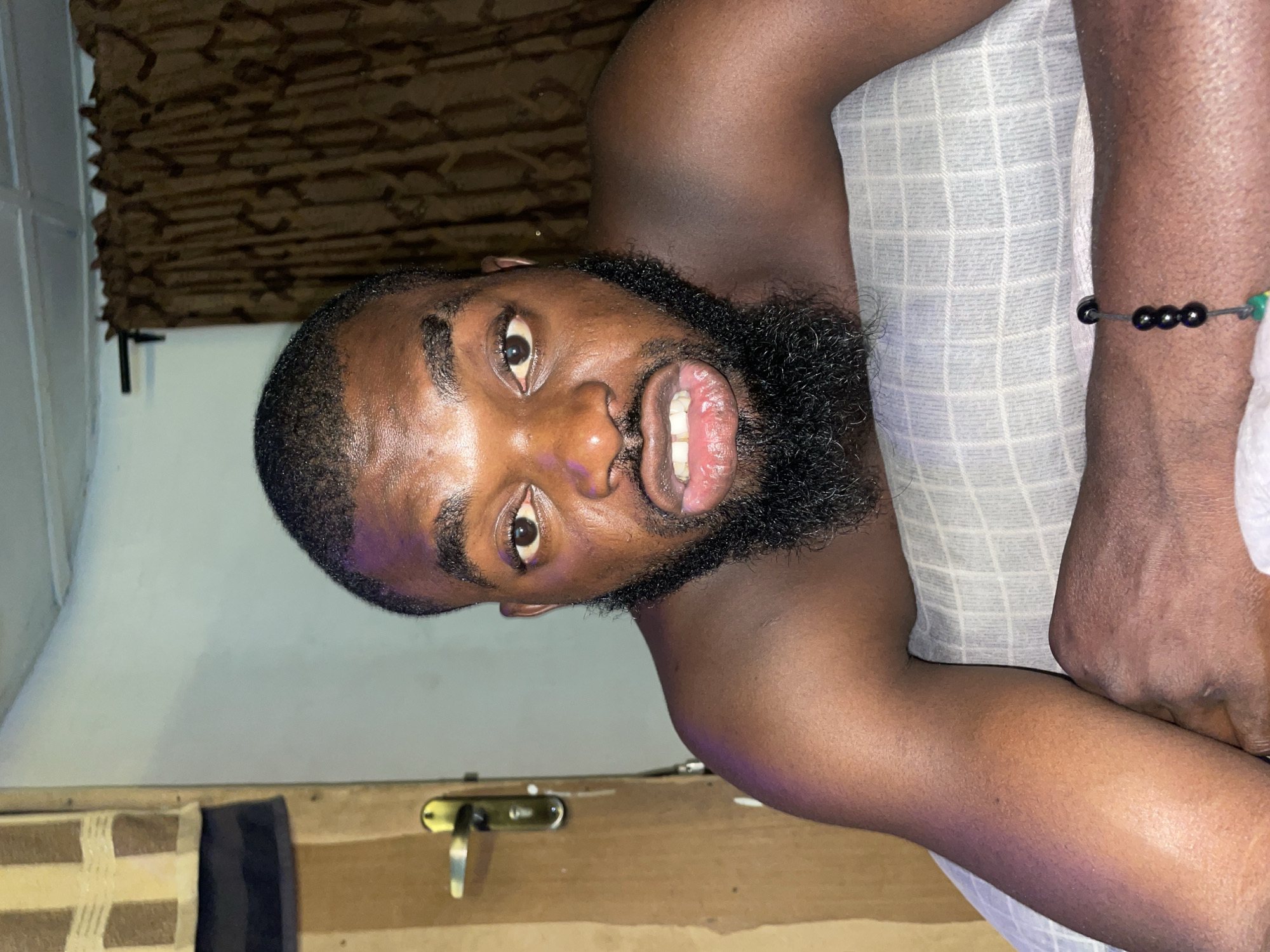 A close-up, eye-level shot shows a Black man lying on his back on a pillow. He has dark skin, a shaved head with short black hair, and a full black beard. His mouth is open, revealing his teeth and gums, and his eyes are wide open, looking directly at the camera. The man is shirtless, and his shoulders and arms are visible.
In the background, a brown patterned curtain hangs behind him. To his left, a wooden door with a gold handle is visible. A section of a patterned towel hangs on the wall to the left of the door. The lighting appears to be artificial, casting a purple hue on the man's skin, particularly on his forehead and shoulder. The setting appears to be indoors, possibly a bedroom, and the overall ambiance is intimate and unposed. There are no visible indications of the city of Jos, Nigeria within the frame.