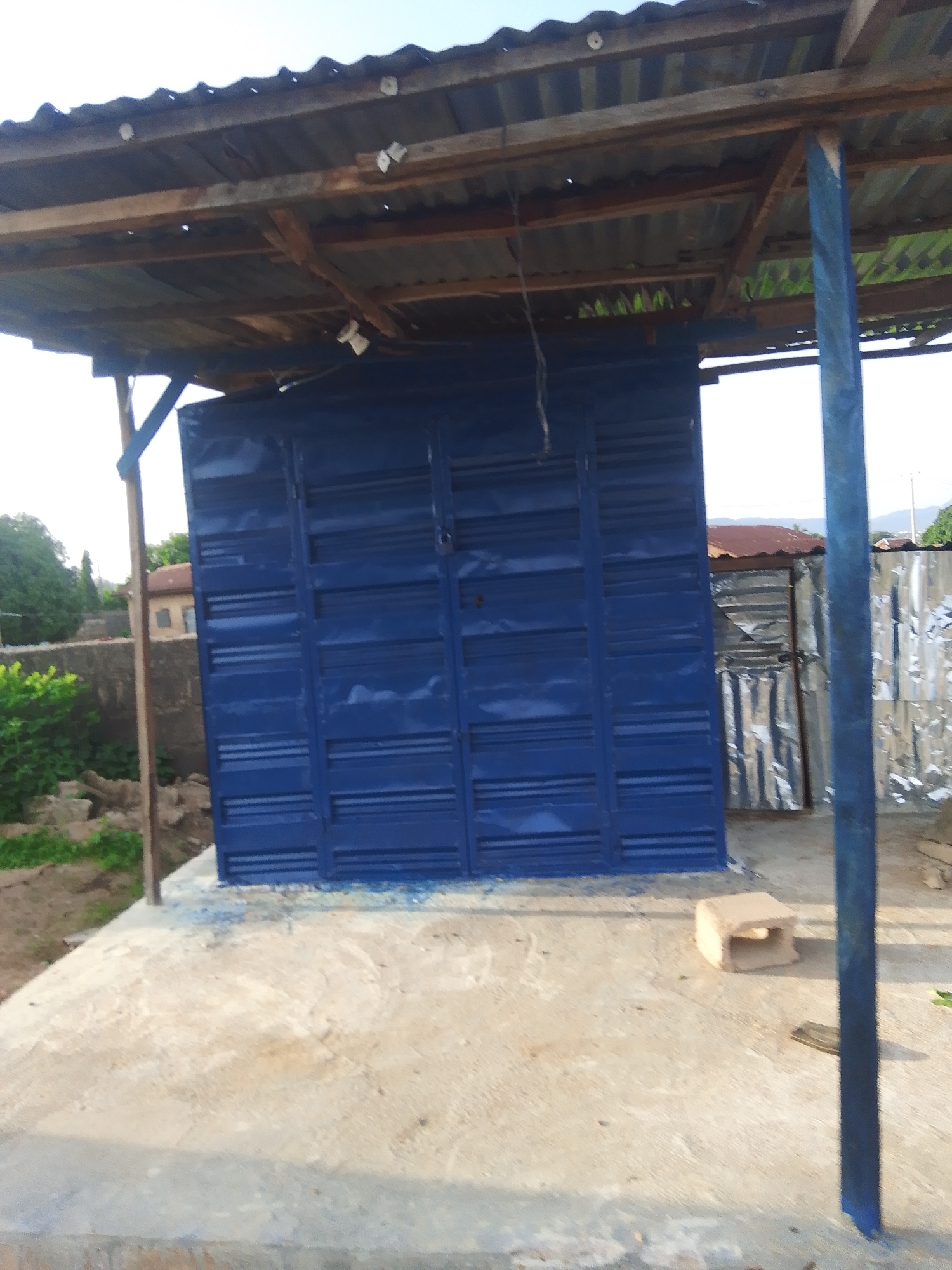 The image depicts an outdoor scene in Jalingo, Nigeria, likely a construction site or an area under development. The central subject is a newly painted, vibrant blue container or structure with a corrugated metal door. This structure is positioned under a rustic, wooden-framed roof covered with corrugated metal sheeting, which shows signs of age and weathering.
To the left of the blue structure, a concrete foundation or platform is visible, with a small, light-colored, hollow brick placed on it. In the background, there are glimpses of other structures, including one with a rusty metal roof and another clad in shiny corrugated metal. Green foliage and trees are present in the middle ground, suggesting a rural or semi-rural setting. A brick wall can be seen behind the bushes. The sky above is clear and bright, indicating it is daytime. The overall impression is one of ongoing construction or the setup of a new establishment. No people are visible in the frame. The scene is bathed in natural light, with shadows cast on the concrete, suggesting it is either morning or afternoon. There is no visible text in the image.