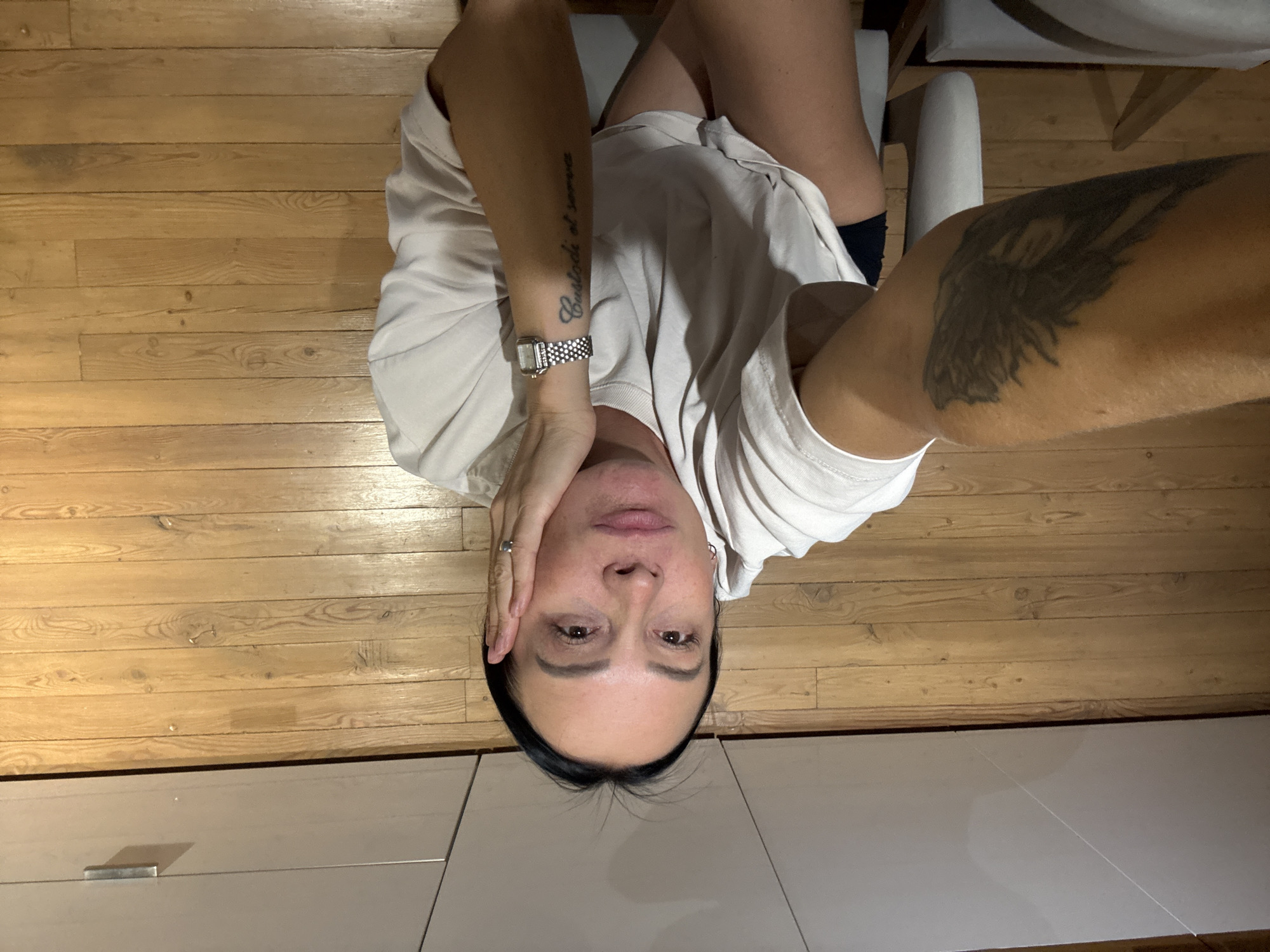 The image shows a woman from the chest up, with her face partially obscured by her hand. She is wearing a light-colored t-shirt and has a watch on her left wrist and a tattoo on her left forearm. Her right forearm, which is in the foreground, also has a tattoo. The background consists of a wooden floor and part of a light-colored cabinet. The image appears to be taken indoors. There is no visible indication of the specific city or location within Chaikovsky, Russia.