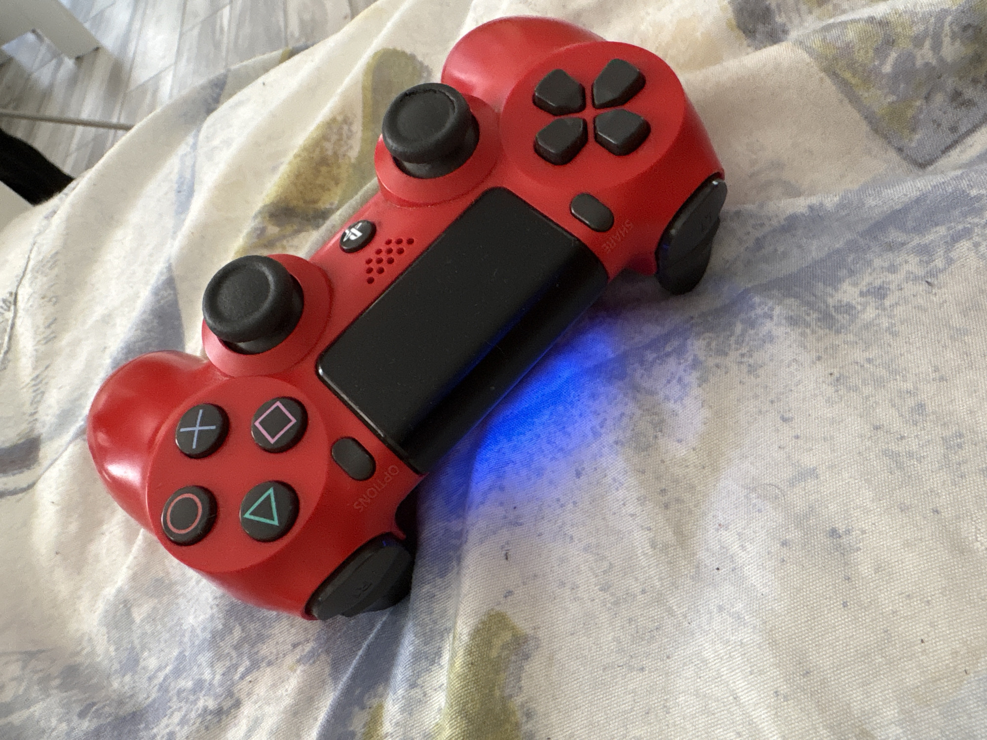 A red PlayStation 4 DualShock controller is lying on a patterned bedsheet with blue, white, and beige colors. A blue light emanates from the controller. The location is not discernible from the image.