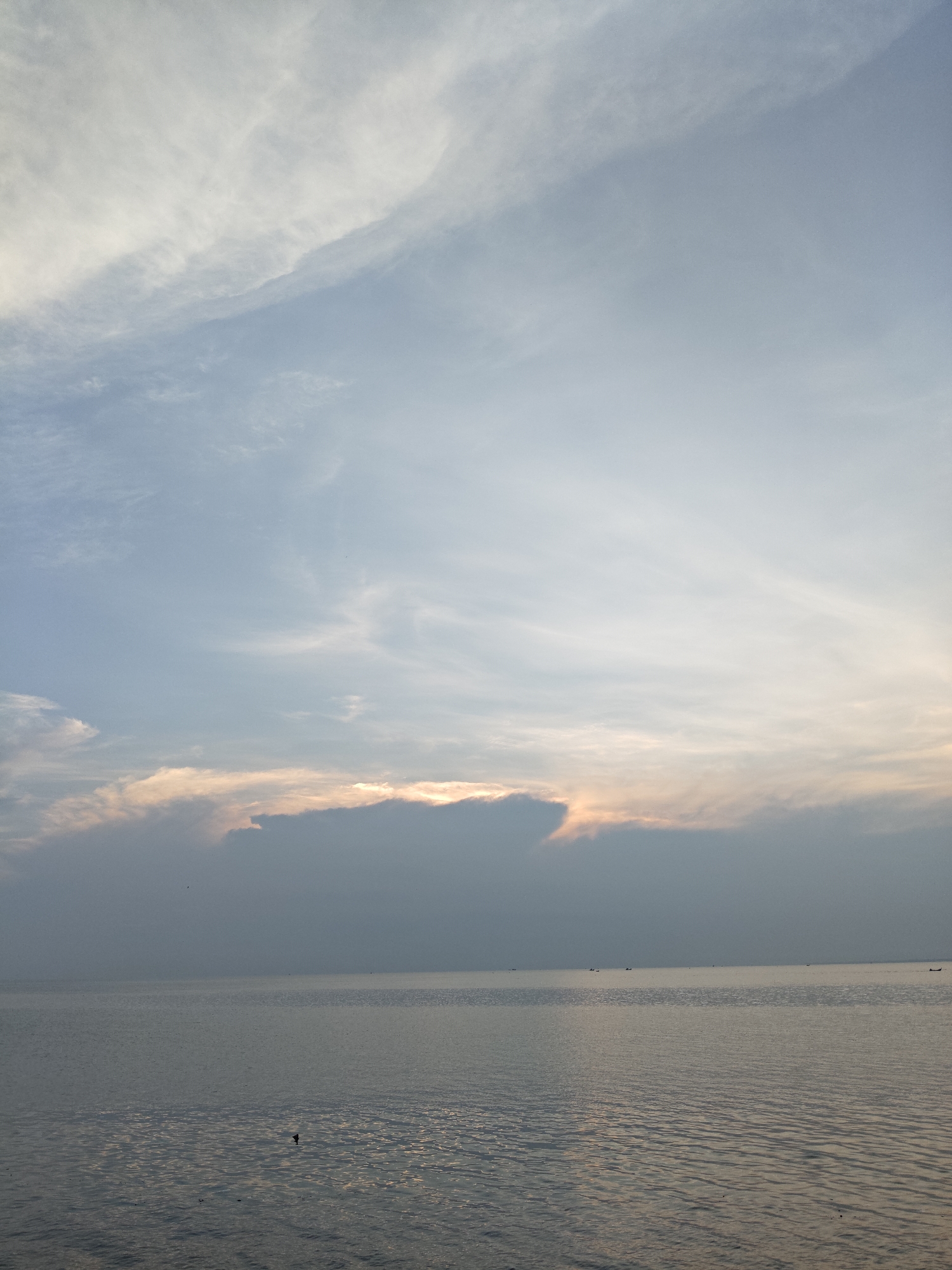 The image depicts a serene seascape. The foreground consists of a calm body of water, its surface reflecting the light, creating a shimmering effect. In the distance, a horizon line separates the water from the sky. The sky is mostly clear with wispy, soft clouds scattered across it. A band of dark clouds hangs low on the horizon, partially obscuring the setting or rising sun, which creates a subtle orange glow around the edges of the clouds. Small boats are visible along the horizon, suggesting human activity. The scene appears to be a coastal area, with a focus on the sky and water. Given the prompt's information, the location is likely in Lhokseumawe, Indonesia.