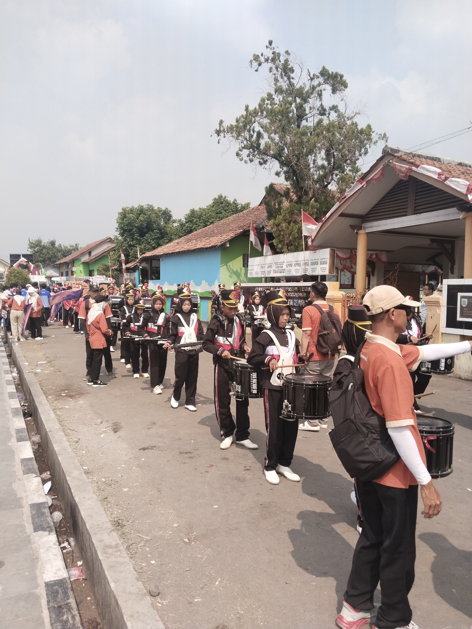 The image depicts a street parade in Margasari, Indonesia. A marching band, composed of individuals in uniform, is moving down a paved street. The band members are primarily wearing black uniforms with white and red accents. Many are carrying drums and drumsticks, suggesting they are actively participating in a musical performance. The scene is set against a backdrop of low-rise buildings, some painted in vibrant colors, and trees. Indonesian flags are visible, indicating a possible national celebration or event. The street itself shows signs of wear and tear, with a concrete gutter running along the side, containing some debris. The sky is bright and overcast, suggesting daytime. Spectators are present on the side of the street, though they are less prominent than the marching band.