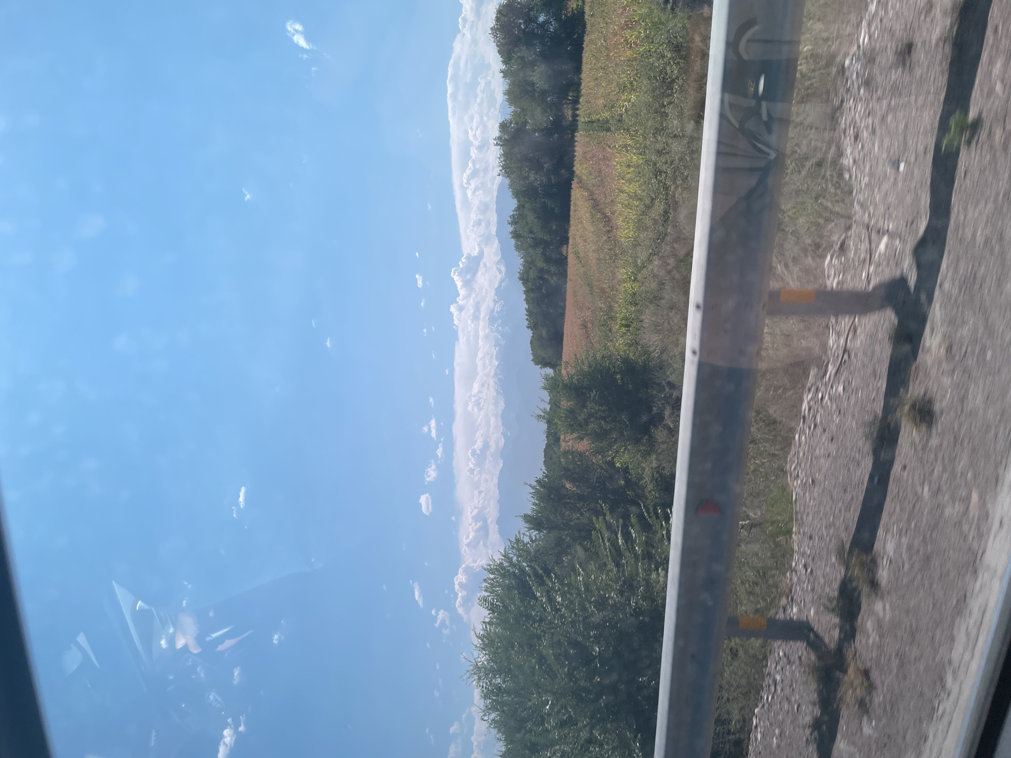 The image captures a scenic view of a rural landscape under a bright blue sky with scattered white clouds. In the foreground, a metallic guardrail runs horizontally across the frame, supported by posts. Beyond the guardrail, a gravelly roadside is visible, with sparse vegetation. The midground features a field of crops, appearing to be corn or a similar grain, with rows of plants stretching into the distance. A line of trees forms a distinct boundary between the field and the background. In the far distance, a range of majestic mountains, possibly snow-capped, dominates the horizon. The overall impression is one of natural beauty and open space, suggesting a location outside of a dense urban environment.