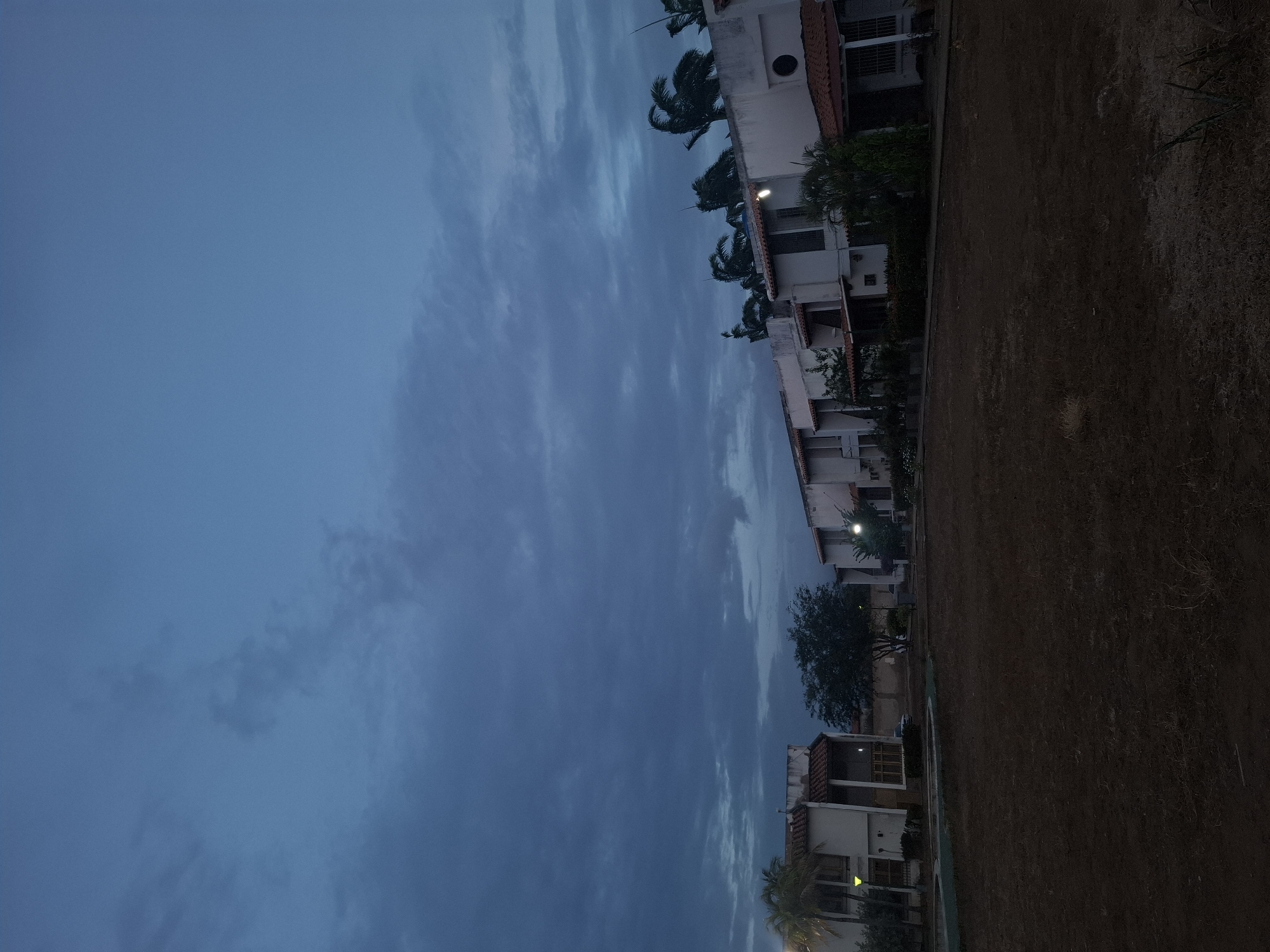 The image depicts a row of residential buildings along the edge of a dry, grassy area, likely a field or park. The buildings appear to be located in Juan Griego, Venezuela, based on the architecture and vegetation. The sky is overcast with dark blue clouds, indicating either dusk or dawn. The buildings are predominantly white with red tile roofs, and some have visible exterior lighting. Palm trees are interspersed among the buildings. The image is taken from a low angle, possibly at ground level. There are no people visible in the scene.
