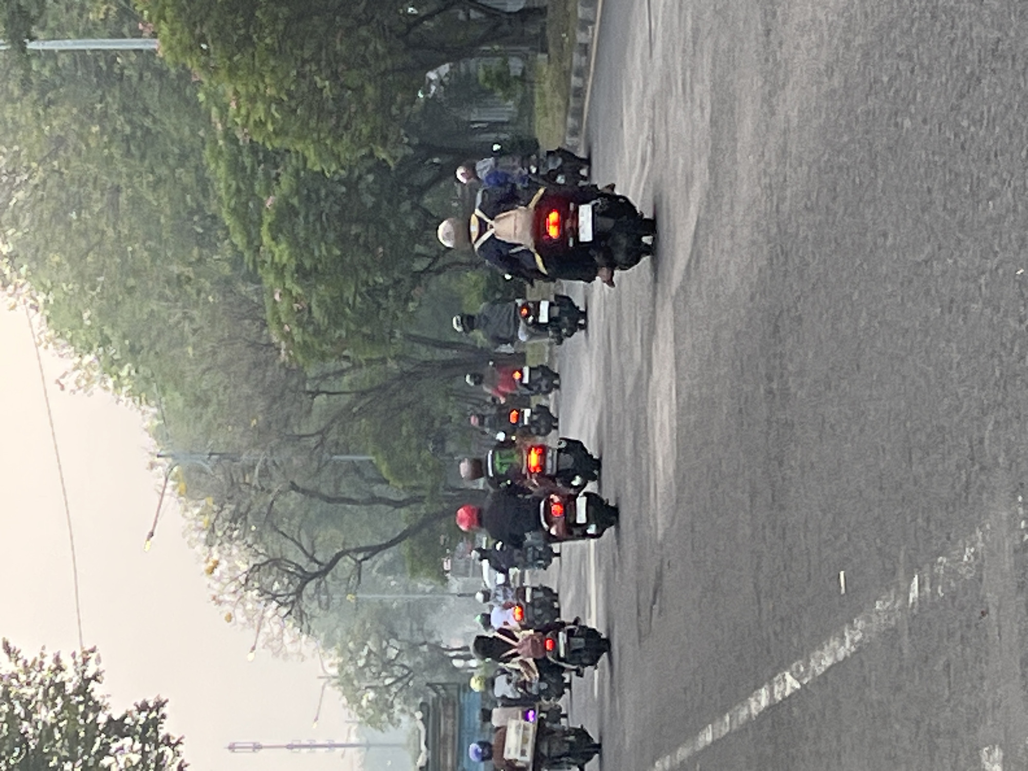 A convoy of motorcycles navigates a paved road lined with trees and streetlights in Gelam, Indonesia. The motorcycles, primarily scooters, are being ridden by individuals wearing helmets, some with backpacks. The road is marked with white lines, and the environment appears overcast, suggesting it could be early morning or a cloudy day. The vehicles are moving in the same direction, indicating a procession or a typical commute. The overall scene depicts a common mode of transportation in the region.