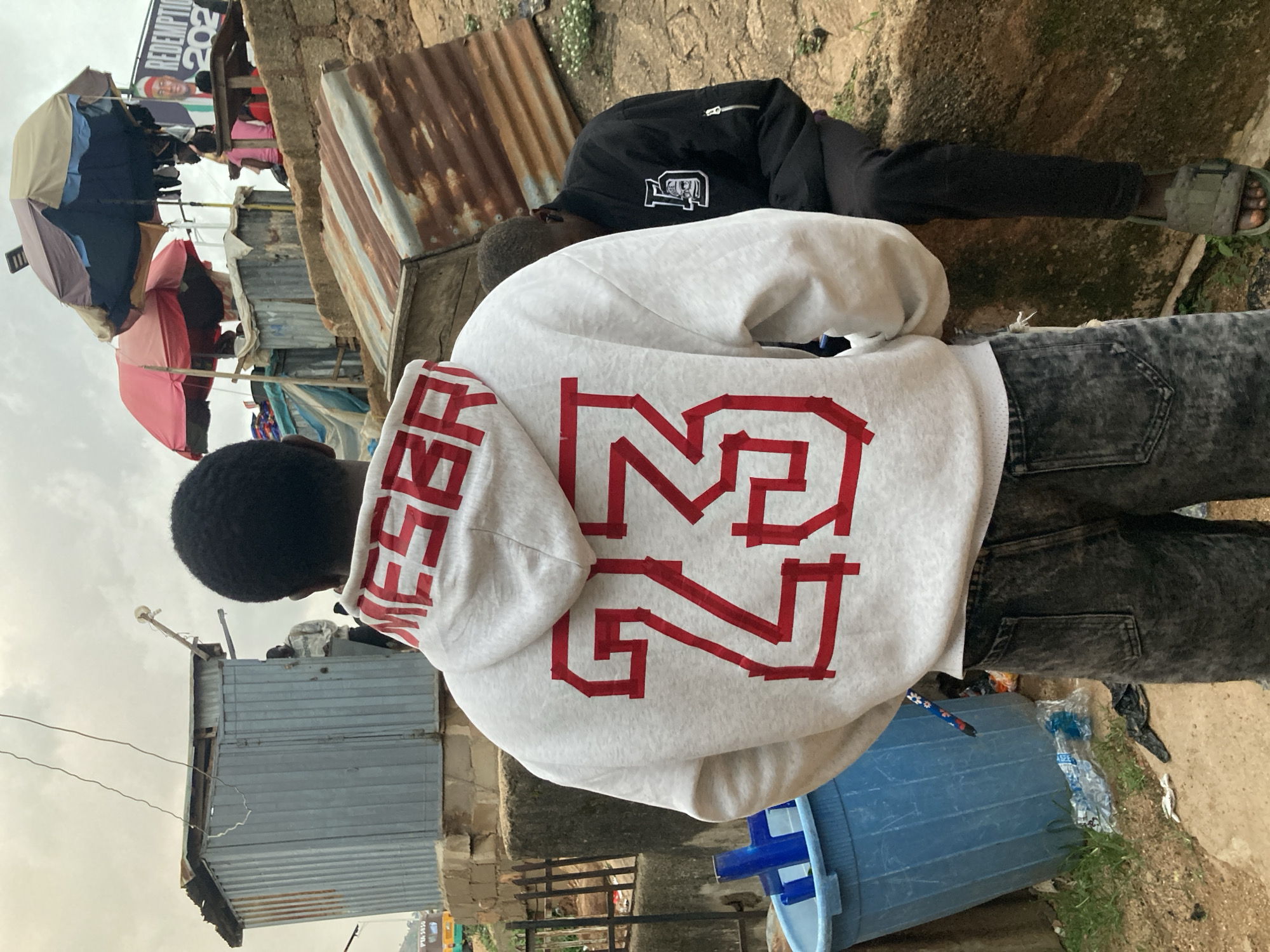 This is a medium shot of a young man from behind, wearing a white hoodie with red lettering that appears to be "MESSI 23". He is standing in what seems to be an informal settlement with corrugated metal structures and some debris on the ground. The ground is dirt and there is a large blue plastic bin. In the background, there are more corrugated metal structures, some covered with tarps or umbrellas, suggesting makeshift shelters or market stalls. A poster with the text "REDEMPTION 2023" is visible in the distance. Another person is visible in the background, wearing a black jacket and dark pants. The lighting suggests it is daytime, possibly overcast. The setting appears to be Gwong, Nigeria, based on the provided location context.