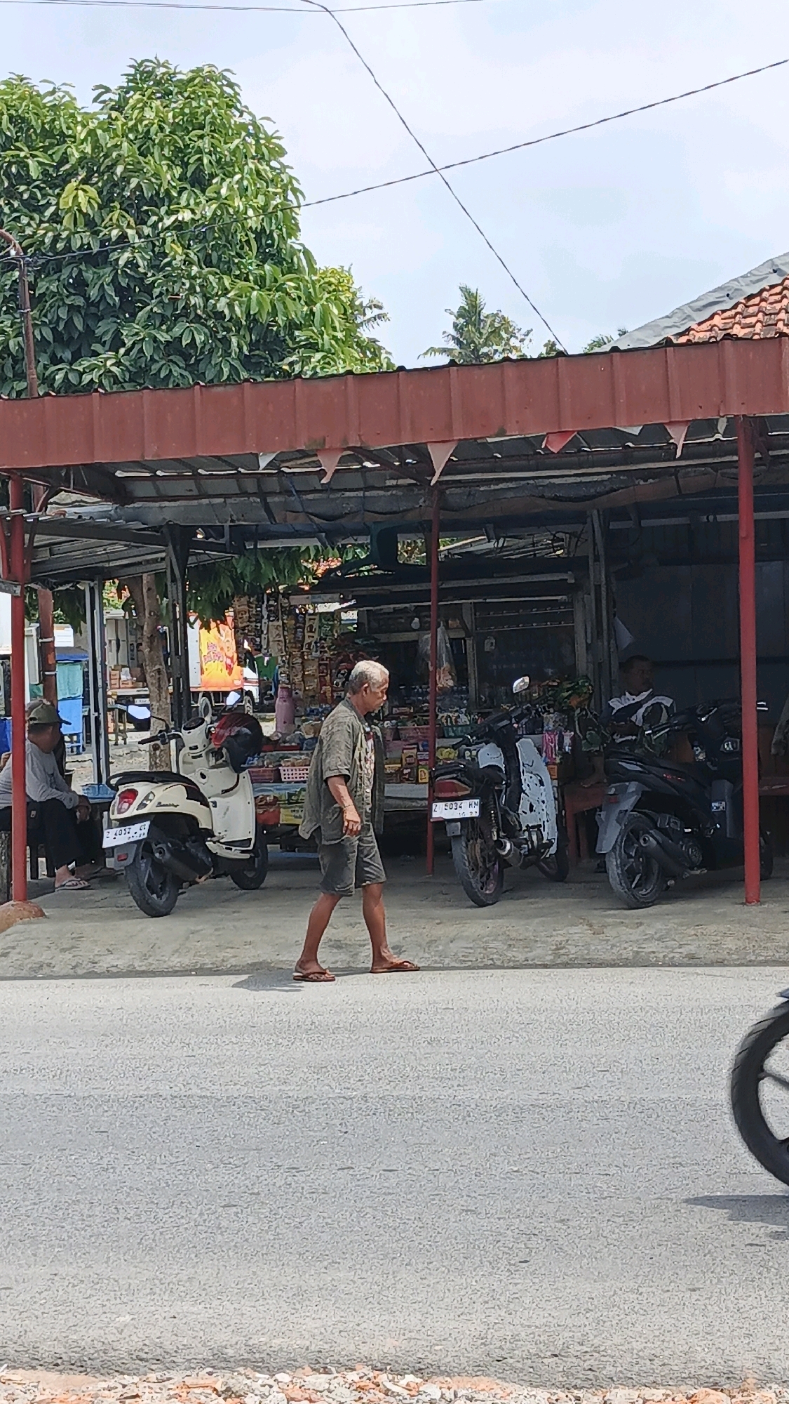 A man walks past a roadside stall in Desa Karangjaladri, Indonesia. The man, an elderly male with gray hair and wearing a green short-sleeved shirt and dark shorts, walks from left to right across the frame. He is barefoot in flip-flops. Behind him, a white scooter is parked. To the right of the scooter, shelves are stocked with various goods, likely merchandise for sale. A red-roofed canopy covers the stall. Two other individuals are visible within the shaded area of the stall. One, wearing a cap and grey long-sleeved shirt, sits on a bench on the left. Another person, wearing a patterned shirt, is seated on the right. Several motorcycles are parked in front of the stall. The scene appears to be along a paved road.