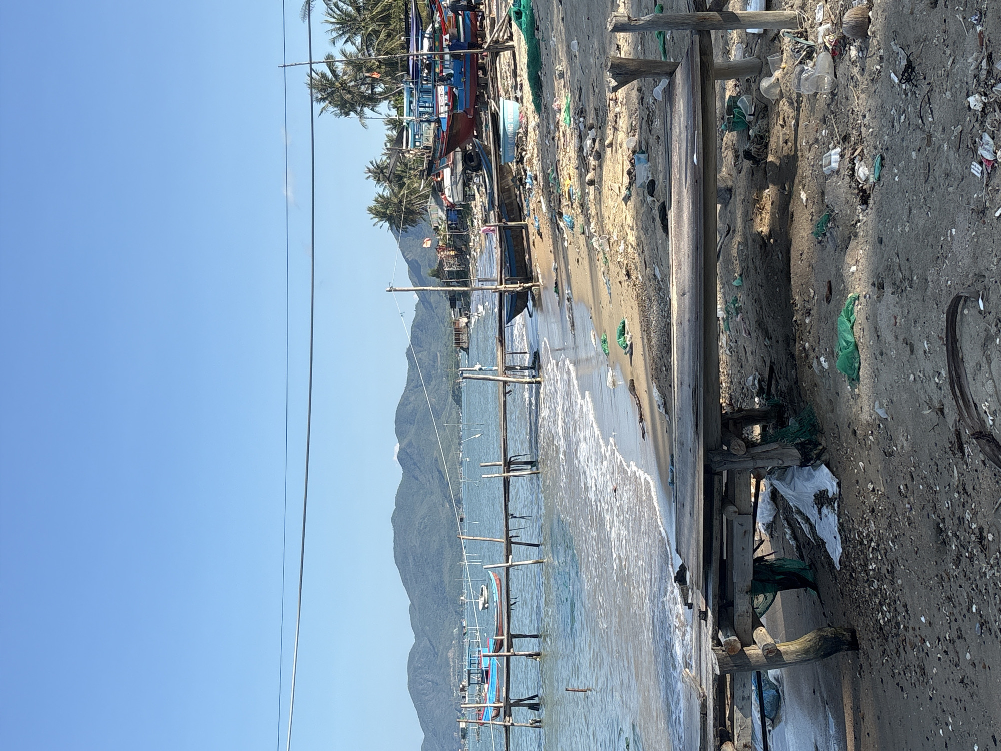 The image depicts a cluttered beach scene in Bac Nha Trang Ward, Vietnam. In the foreground, a rickety wooden structure runs parallel to the shoreline, positioned between the sandy beach and the water's edge. The beach is littered with trash, including plastic bags, cups, and other debris. Numerous fishing boats are moored just offshore, and more boats and fishing nets are present along the beach, adjacent to a building. In the background, mountains are visible, partially obscured by the clear blue sky. Power lines cross the sky. No people are apparent in the view.