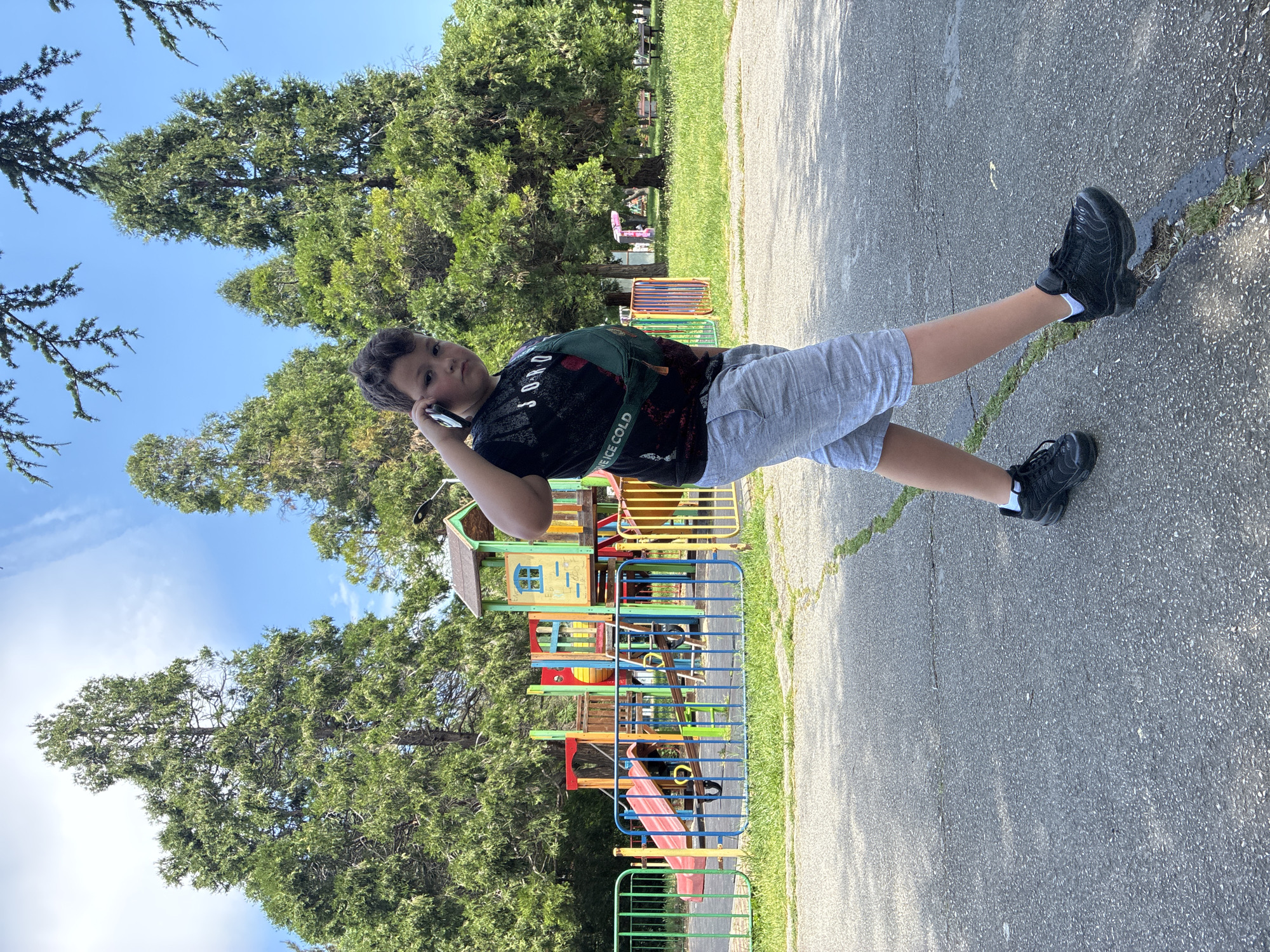 A boy stands in a park near a playground in Varna, Bulgaria. He is talking on a cell phone. The playground has a slide and climbing structure. The park has trees and paved walkways.