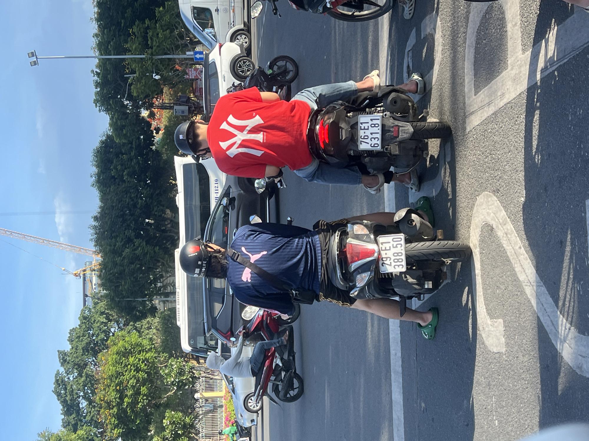This is a photograph taken in Đà Nẵng, Vietnam, likely in a busy urban area with traffic. The image focuses on two individuals riding motorbikes side-by-side, facing away from the camera. The person on the left is wearing a dark blue long-sleeved shirt with a pink Puma logo and black shorts, riding a red motorbike. They have green Crocs on their feet. The person on the right is wearing a red t-shirt with a white New York Yankees logo and blue jeans, riding a black motorbike. They are wearing white sandals. Both individuals have helmets on. The motorbikes have license plates visible: the one on the left reads "29-E1 388.50" and the one on the right reads "76-E1 631.81".
In the background, other vehicles are present, including a white van with a sign that appears to have Vietnamese text and numbers, a silver car, and another white van. Trees line the street, and a construction crane is visible in the distance, suggesting ongoing development. The sky is blue with some clouds. The road surface is asphalt with white painted lane markings. The lighting suggests it is daytime, possibly late afternoon due to the shadows. The scene captures a common mode of transportation and daily life in a Vietnamese city.