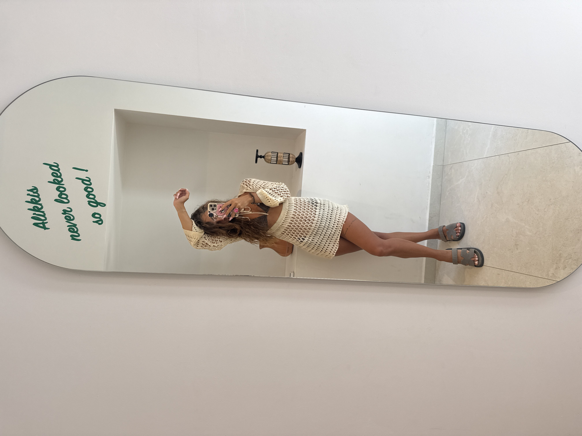 A person is taking a selfie in a full-length arched mirror. The mirror reflects a woman with long, wavy brown hair, wearing a crocheted cream-colored top and skirt set over a brown bikini. She is posing with one arm raised above her head and holding a pink-cased smartphone in her other hand. The mirror has green text written across the top that reads "Alikkis never looked so good!".
The reflection also shows a white wall with an alcove containing a decorative wicker object. The floor is tiled with a light beige and white marble pattern. The environment suggests a domestic or hospitality setting, likely indoors. The text on the mirror refers to "Alikkis," which is likely a place or a person's name associated with Stavros, Greece, as implied by the prompt.