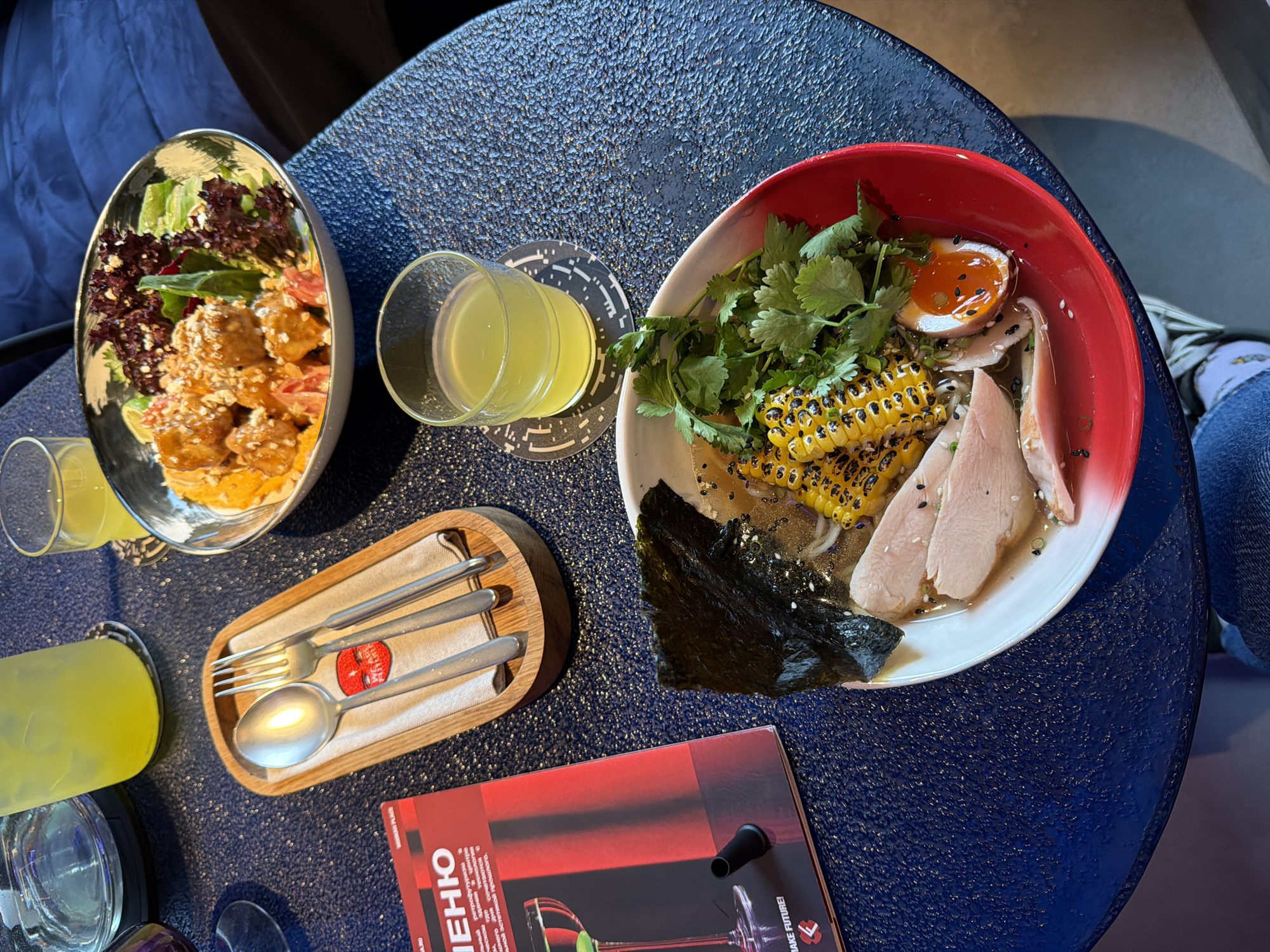 The overhead shot shows a dark blue, textured table in a restaurant. On the table are two bowls of food and two glasses of yellow liquid. One bowl contains ramen with chicken, corn, a soft-boiled egg, and seaweed. The other bowl holds a salad with what appears to be fried chicken pieces and lettuce. One glass of yellow liquid is on a coaster with a geometric pattern. Next to the ramen bowl is a red menu with text in Russian, reading "МЕНЮ". In the upper left corner, a large glass of yellow liquid is visible. A wooden tray with cutlery, including a spoon and fork, rests on a white napkin. The setting appears to be a modern restaurant, likely in Moscow, Russia, given the Russian text on the menu. No people are clearly visible, but a person's knee and jeans are in the lower part of the image.