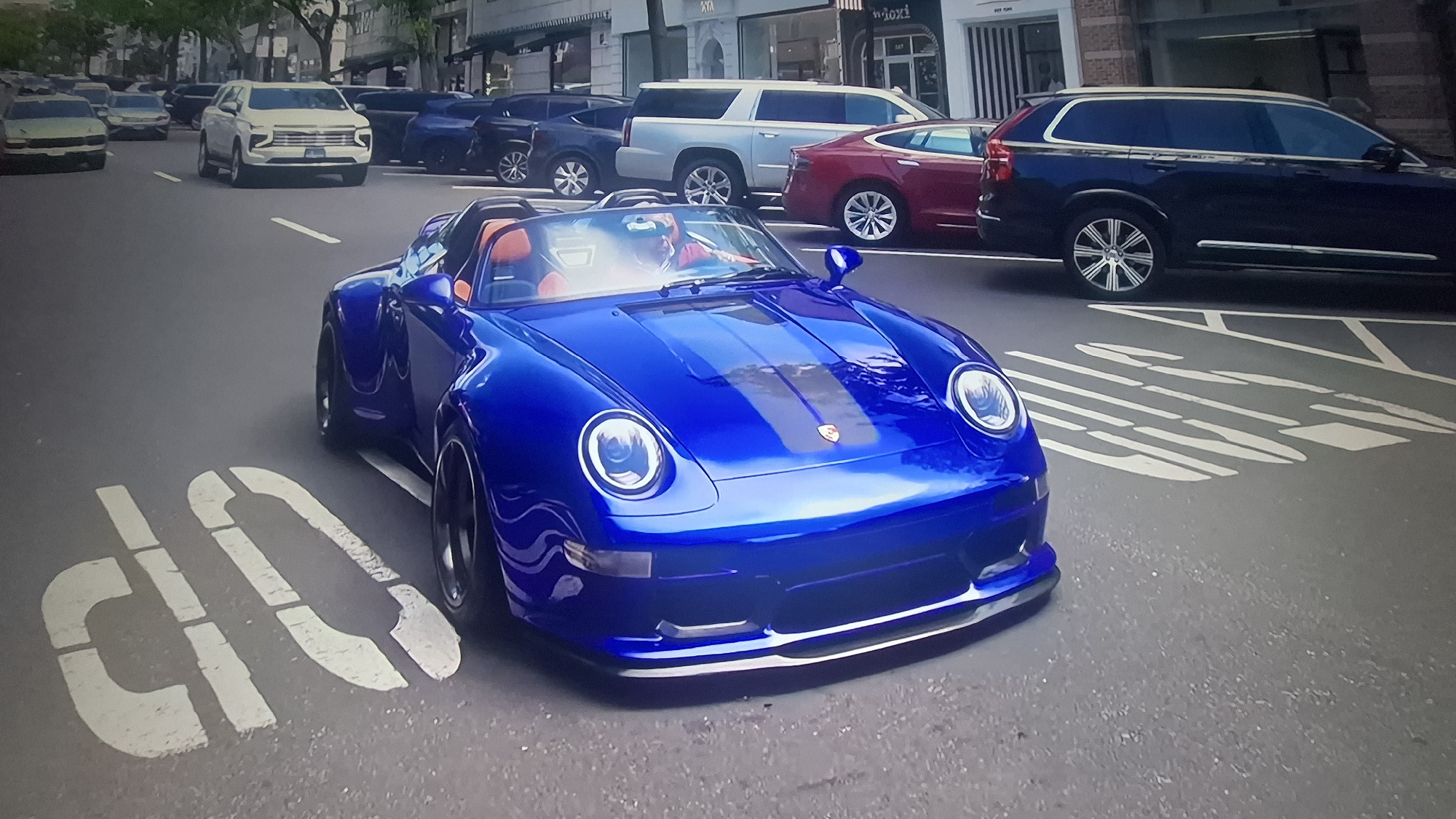 The image shows a bright blue Porsche 959 Speedster parked on a city street, facing the camera. It is positioned on an area marked with white painted "DO NOT STOP" text on the asphalt. Several other vehicles are parked behind the Porsche along the street, including an off-white SUV, a dark red sedan, and a dark blue SUV, as well as some other cars visible further down the street. The location appears to be an urban shopping area, based on the building facades and storefronts visible in the background. There is a driver visible in the car. The overall lighting suggests it is daytime.