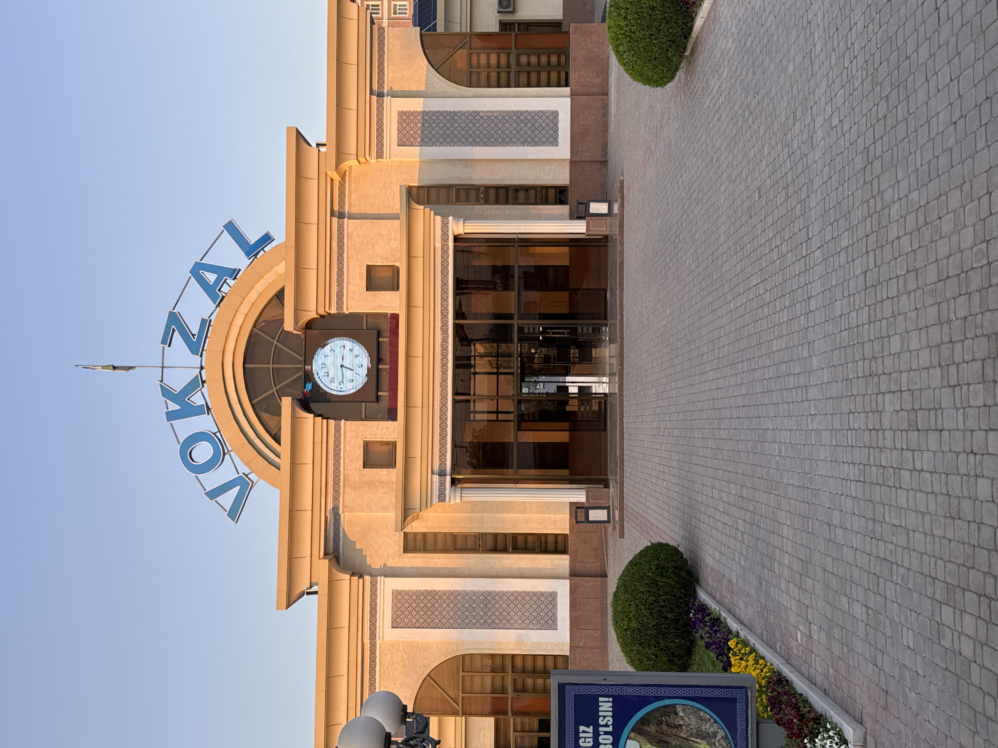 The image depicts the facade of the Jizzakh railway station in Uzbekistan. The building features a clock tower with a large analog clock displaying the time. Above the clock, the Cyrillic letters "VOKZAL" are visible, spelling out "вокзал," which means "railway station" in Russian. The architectural style appears to be a mix of Soviet-era and Central Asian influences. The entrance is marked by glass doors and decorative elements. The surrounding area includes paved walkways and ornamental landscaping with flowers and shrubs. A blue sign with Cyrillic text is visible near the bottom of the frame. The overall lighting suggests it is either early morning or late afternoon.