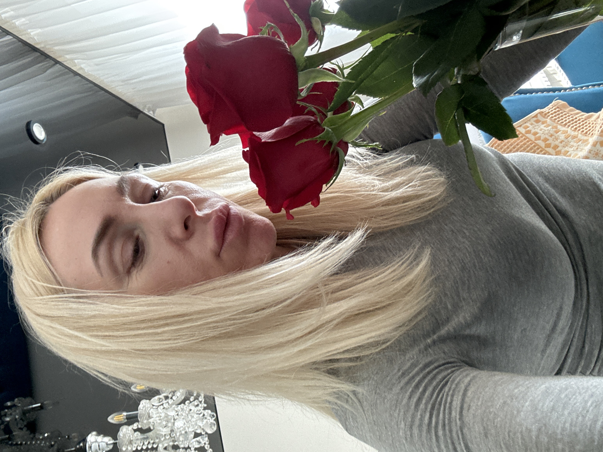 A close-up, high-angle shot captures a woman holding or positioned near a bouquet of red roses indoors. The woman, with long blonde hair, is partially visible, primarily her face in profile and her upper body. She wears a grey long-sleeved top. Her expression appears neutral or slightly contemplative as she looks towards the left side of the frame.
The red roses are a prominent feature, with several blooms and green foliage visible in the upper right portion of the image. The setting appears to be a room, indicated by various background elements. To the left, a dark, reflective surface, possibly furniture, is present, along with a circular light fixture and a decorative, crystal-like chandelier. To the right, a bright blue upholstered piece of furniture, possibly a sofa or chair, is visible, along with a textured, light-colored fabric, which could be a blanket or cushion. The ceiling or a white curtain is seen at the top of the frame. The overall lighting suggests an indoor environment, possibly during the day. There is no visible text.