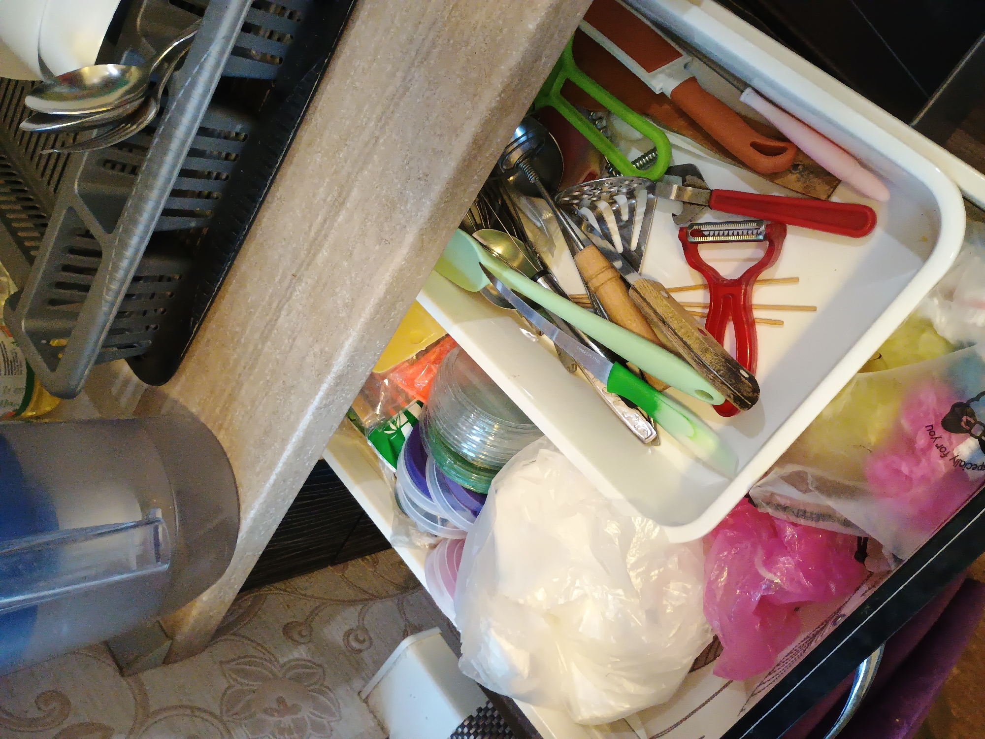 The image shows an open kitchen drawer filled with various utensils and kitchenware. In the foreground, on the left, a dish drying rack holds several metal spoons and forks. The drawer itself is white and contains a variety of items including a red peeler, a potato masher with a wooden handle, green silicone spatulas, and a small knife with a green handle. There are also plastic containers and bags with pink and white contents, possibly food storage bags or packaging. The background is a blurred view of a kitchen counter and floor. The scene appears to be indoors, likely a kitchen, with artificial lighting. There is no visible text that can be clearly read. The overall impression is one of a cluttered but functional kitchen drawer.