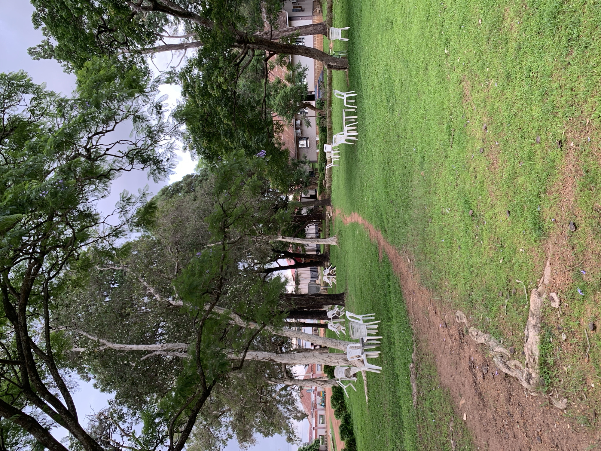 This image depicts an outdoor seating area, likely part of a guesthouse or hotel in Jos, Nigeria, surrounded by lush greenery and mature trees. Several white plastic tables and chairs are arranged on a well-maintained lawn, suggesting a space for guests to relax or dine. The ground is a mix of grass and reddish-brown soil, with exposed tree roots visible near a dirt path that winds through the scene. In the background, a low-rise building with a corrugated roof can be seen, hinting at accommodation facilities. The overall atmosphere is serene and natural, with the dense foliage providing shade.
