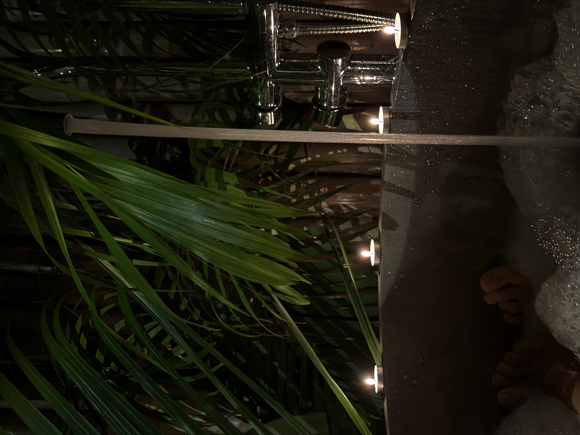In Baan Bo Phut, Thailand, a bathtub filled with foamy water is illuminated by the warm glow of tea lights. The scene suggests a tranquil atmosphere, possibly within a resort or private residence, given the presence of lush green foliage framing the bathtub. The water is flowing from a modern chrome faucet, indicating it's either being drawn or the bath is in use. Bare feet, partially submerged in the bubbles, are visible at the bottom of the tub, hinting at someone enjoying a relaxing soak. The overall impression is one of calm and indulgence, enhanced by the dim lighting and the tropical greenery.