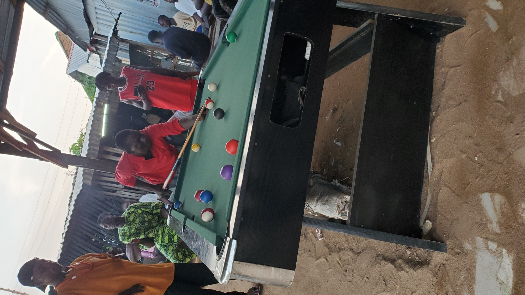 In Jalingo, Nigeria, a group of individuals are gathered around a pool table situated outdoors. The scene appears to be in a semi-urban or rural setting, with corrugated metal roofing and wooden structures forming the background. The ground is sandy and uneven.
The central focus is a pool table with green felt, on which several billiard balls are arranged. A man in a red long-sleeved shirt is actively playing pool, holding a cue stick and aiming at a ball. He is bent over the table, engaged in the game.
To his left, another man wearing an orange t-shirt and a black baseball cap stands watching. Further to the left, a man in a green patterned traditional garment is seated, also observing the game.
In the background, several other people are visible, some seated, others standing, suggesting a social gathering or a casual sporting event. One man in the background wears a red basketball jersey with the number 23. The overall atmosphere seems relaxed and informal. The lighting suggests it is daytime, possibly late afternoon.