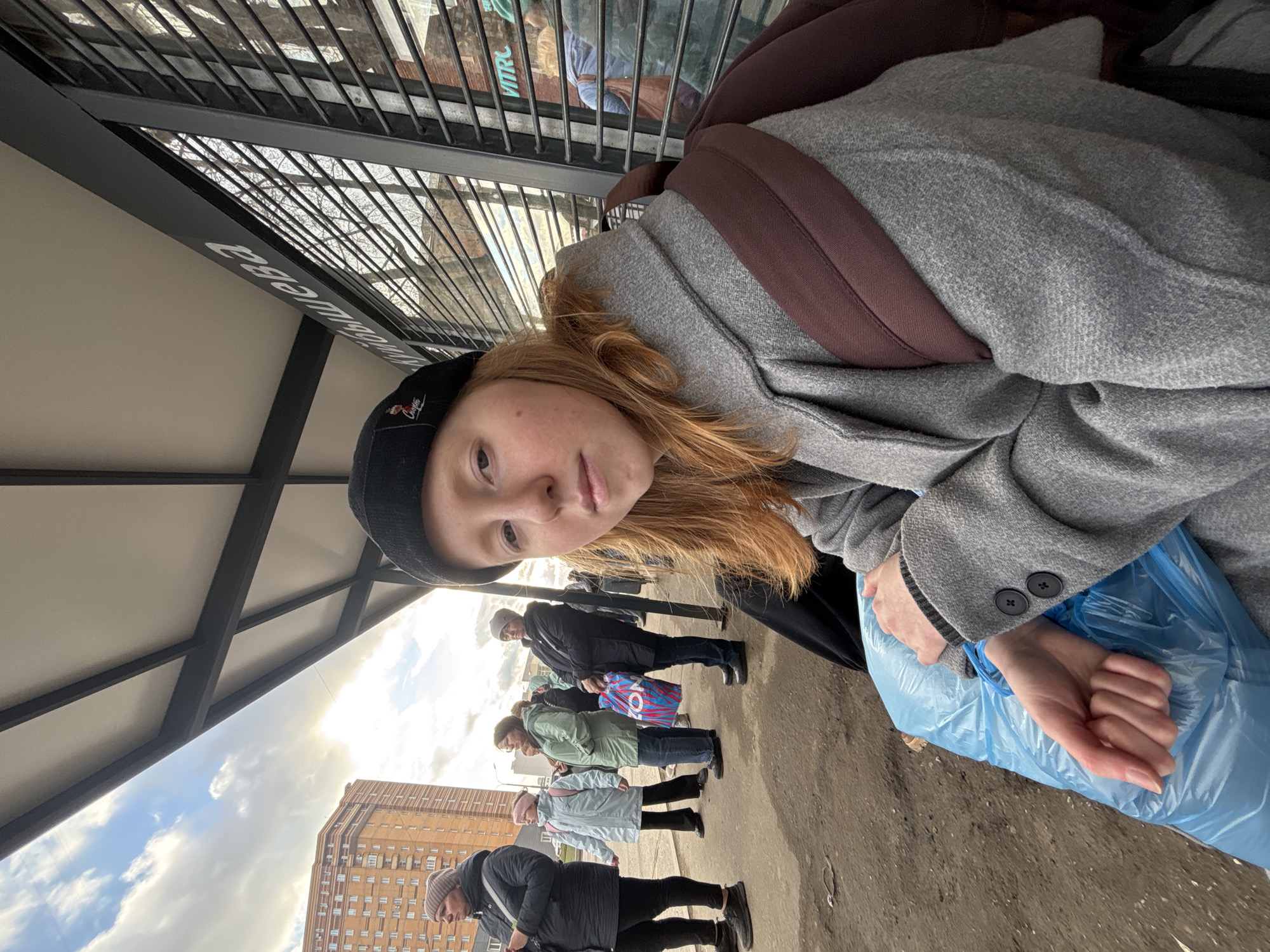 A young, light-skinned person with reddish hair, wearing a black beanie, a gray coat, and a brown backpack, is holding a blue plastic bag. The individual is standing under a bus stop structure with a black metal frame and a white roof. Behind them, a line of people stands on a gray concrete sidewalk. In the background, there is a tall, brown brick building with numerous windows and a cloudy sky. The bus stop sign in Russian reads "кинотеатр мира". This translates to "Mira Cinema". This location is in Perm, Russia.