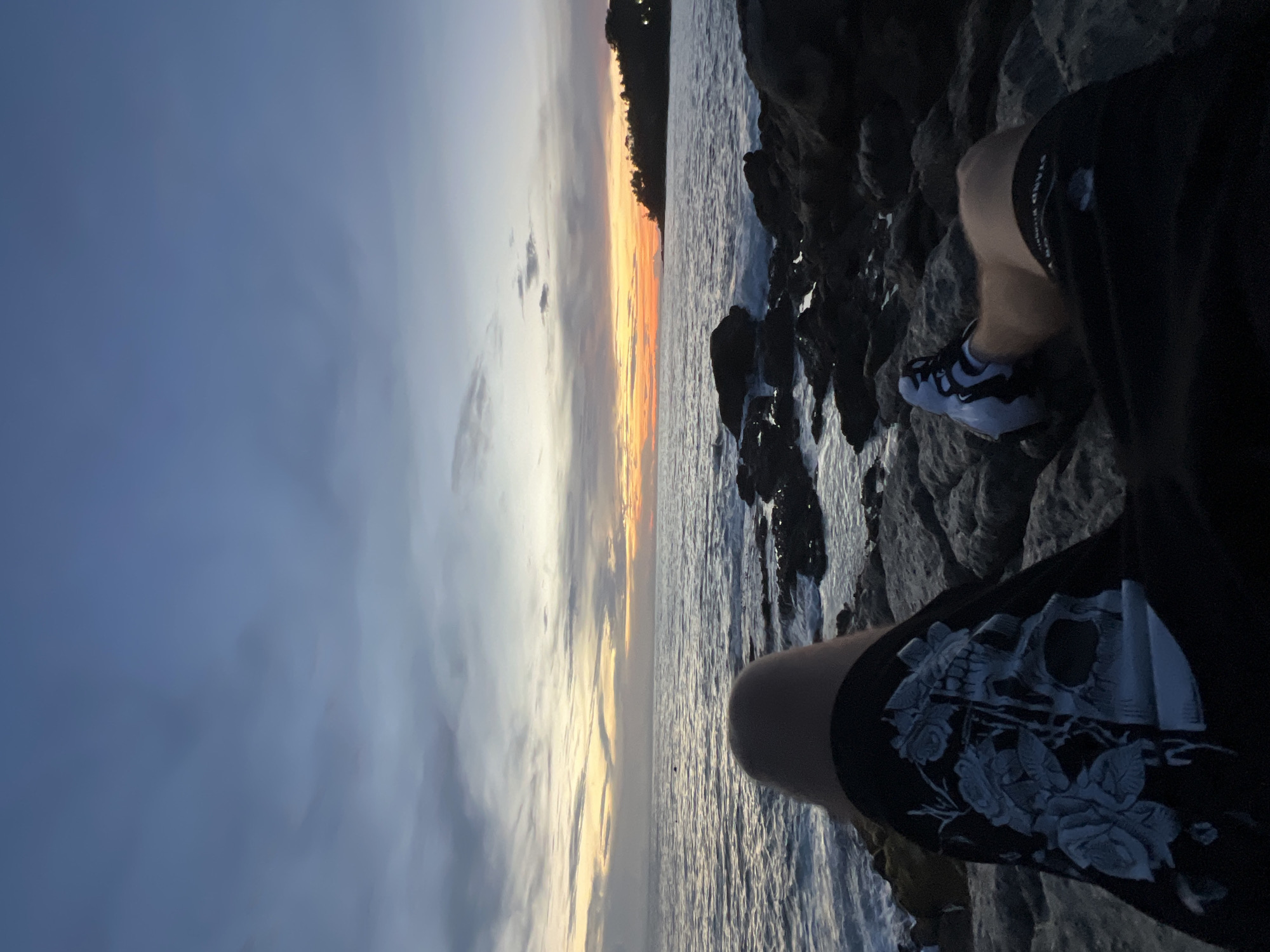 A person is sitting on rocks near the ocean as the sun sets. The sky is filled with clouds and streaks of orange and yellow from the setting sun. The ocean is calm, with small waves breaking against the rocks. The person is wearing shorts with a skull and rose design, and sneakers. The location is on the coast of Choeng Thale, Thailand.