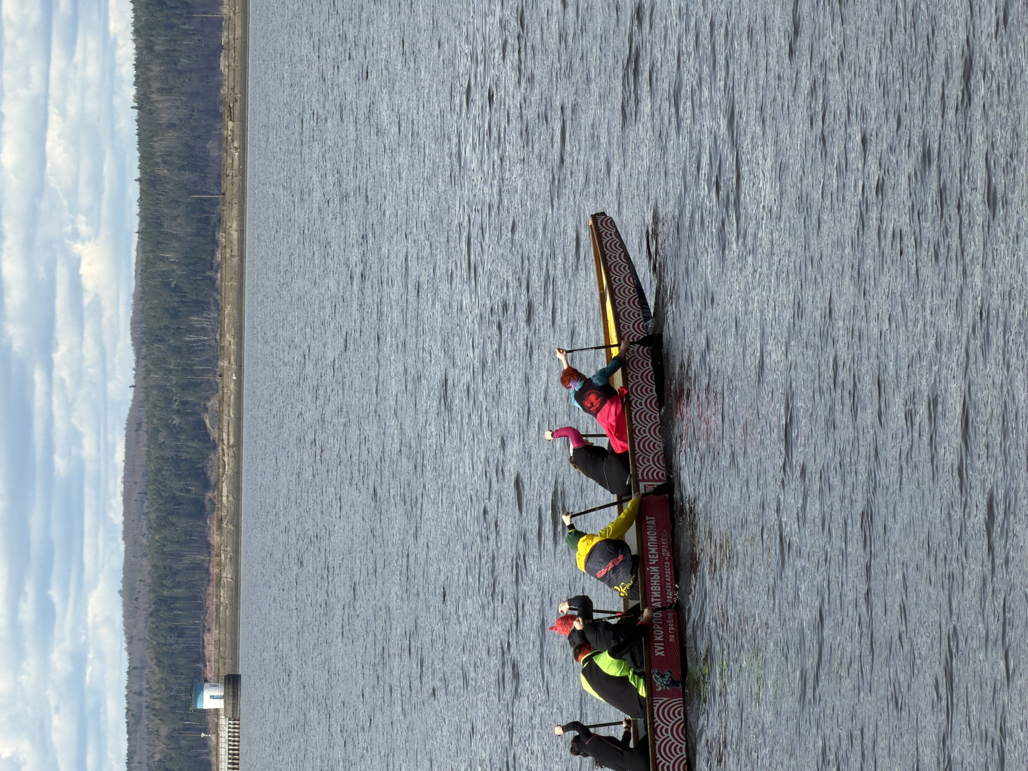 A dragon boat with several paddlers is prominently featured on a calm body of water. The boat is long and narrow, adorned with a decorative red and white wave-like pattern along its side. Clearly visible text in Russian on the boat reads: "XVI КОРПОРАТИВНЫЙ ЧЕМПИОНАТ" (XVI CORPORATE CHAMPIONSHIP) and "по гребле на лодках класса «ДРАКОН»" (in dragon boat racing), indicating a competitive event.
Six individuals are visible within the boat, each holding a paddle and seated in a paddling stance, suggesting they are actively engaged in or preparing for racing. They wear a variety of sportswear, including bright colors such as pink, yellow, and red. Their focused postures convey concentration on the activity.
The setting is a large expanse of water, showing minor ripples on its surface under bright daylight. In the distant background, a forested shoreline stretches horizontally, densely covered with trees. A distinct embankment or wall runs along the water's edge on this distant shore. Towards the left, a small, light-colored building or structure can be seen near the water on the shore. The sky above the treeline is partly cloudy, with visible patches of blue. The weather appears fair and calm. This scene takes place in Chaikovsky, Russia.