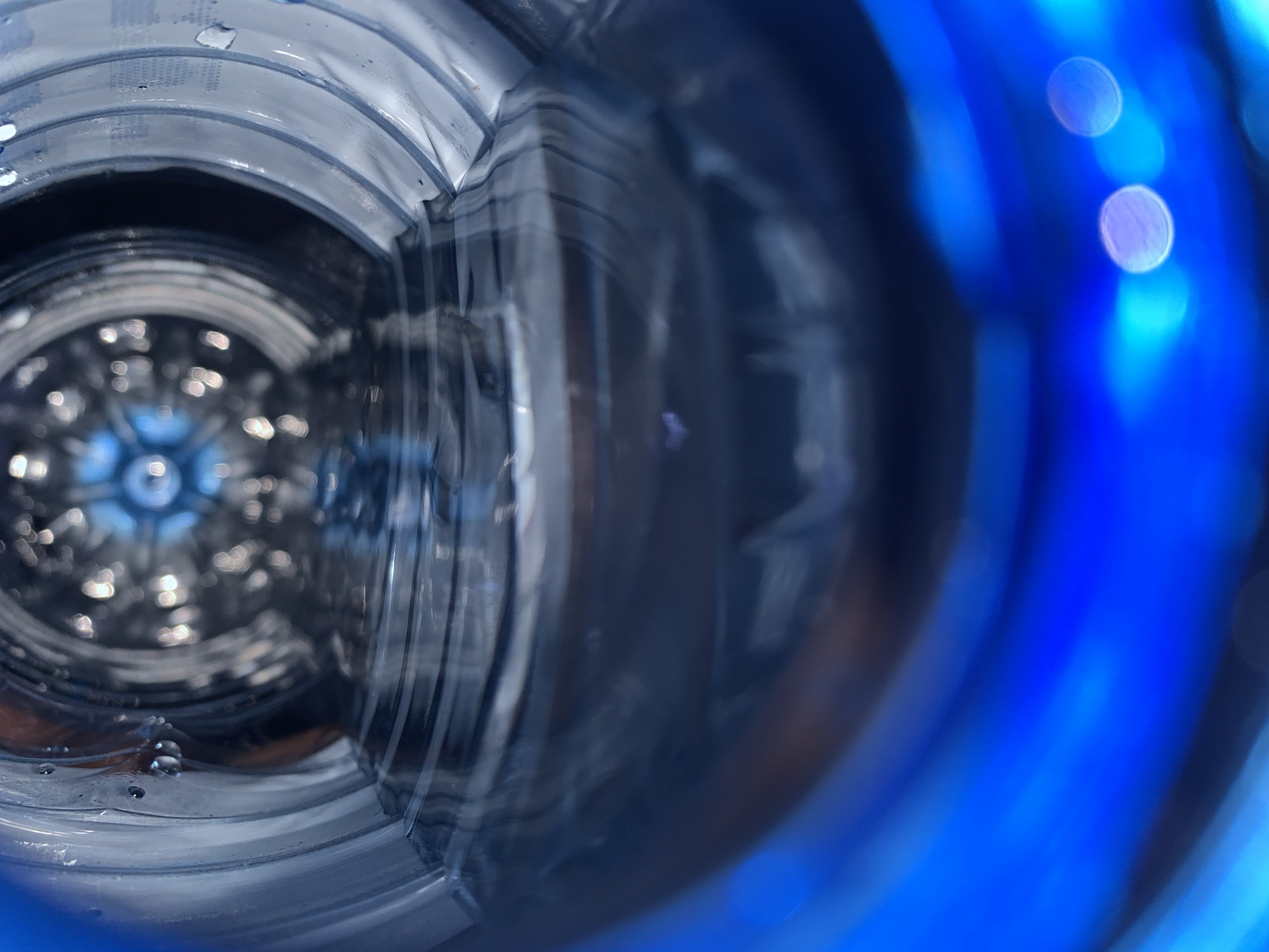 The image appears to be a close-up view looking inside a blue plastic bottle. The interior of the bottle is visible, showing its ridged structure and the bottom, which has a star-shaped pattern. There are some droplets of liquid clinging to the inner surface of the bottle. The focus is blurry, especially towards the foreground where a bright blue section of the bottle is visible, with bokeh lights. There is no discernible location, people, subjects, actions, or interactions. The environment is the inside of a plastic bottle.