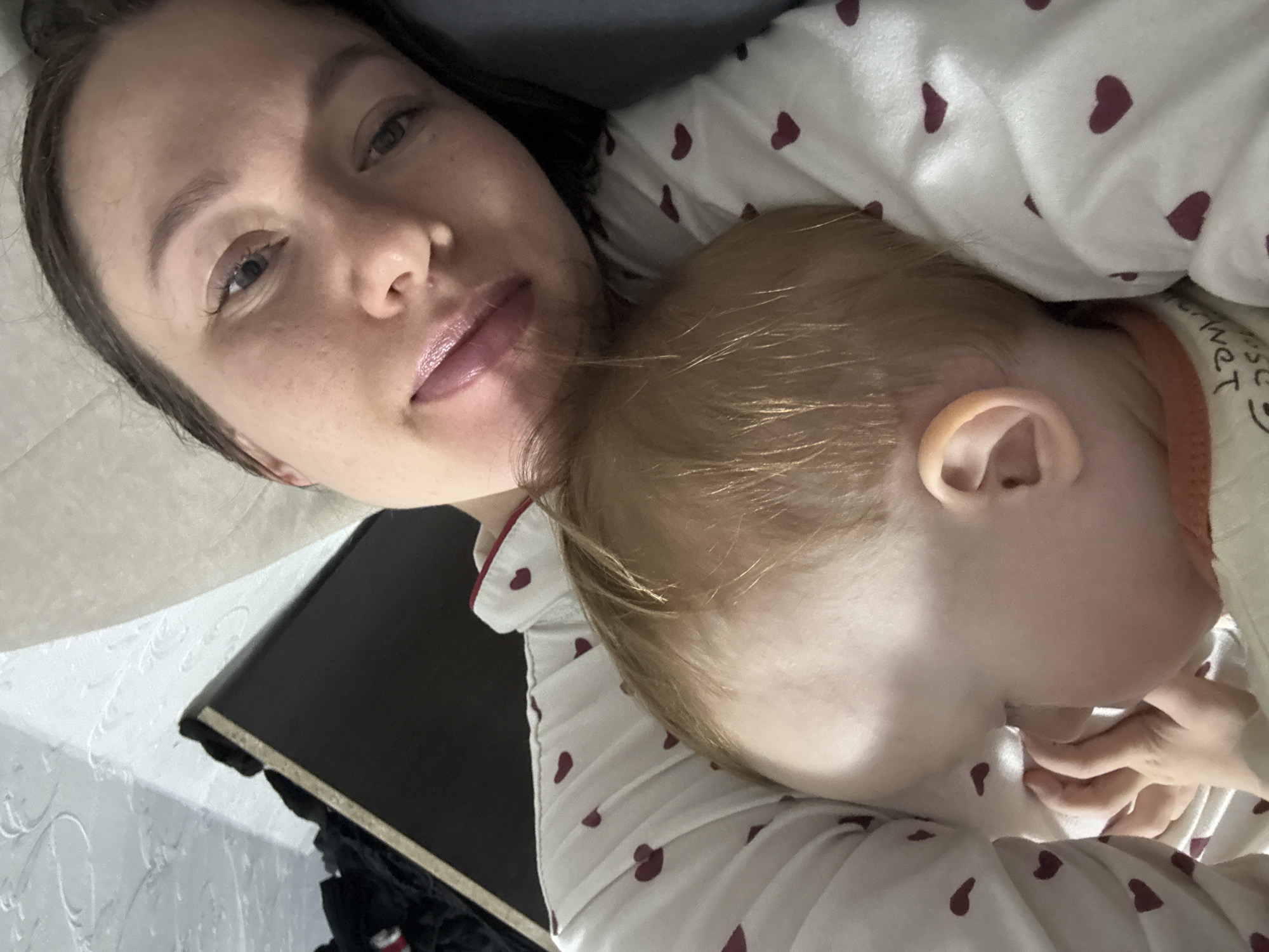 A woman and a baby are lying down. The woman is fair-skinned with brown hair. The baby has blonde hair. They are wearing matching pajamas with a heart pattern. The photo may have been taken indoors, possibly in a residential setting. No specific location in Ферма, Russia, can be identified from the image.