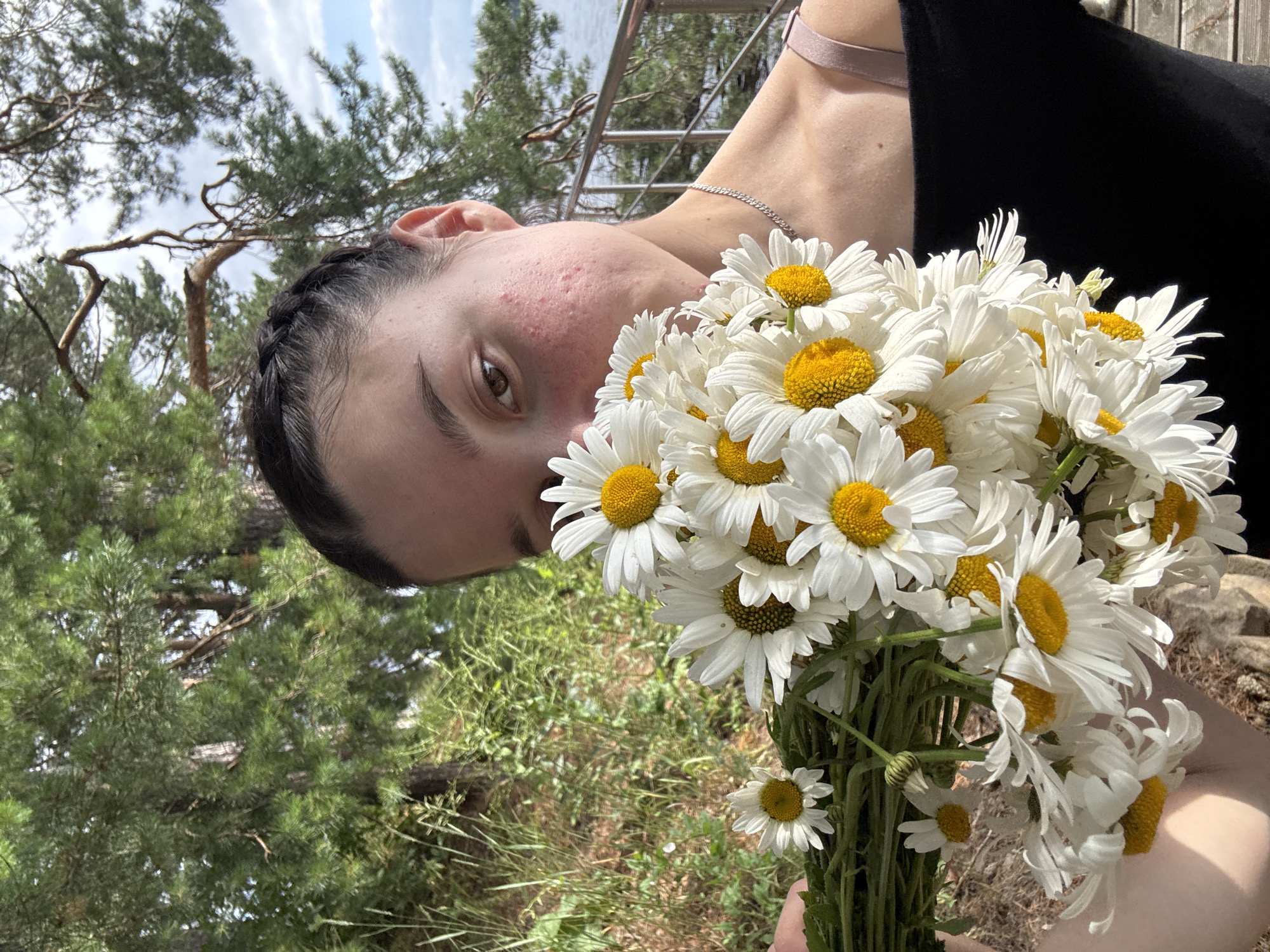 A young woman with dark hair braided to the side is holding a bouquet of white daisies in front of her face. She is outdoors, with trees and foliage in the background. To the right, a body of water and a railing are visible, suggesting a location near a lake or river. The woman is wearing a black off-the-shoulder top. The lighting suggests it is daytime. The scene appears to be in or around the village of Pozdyshki, Russia.